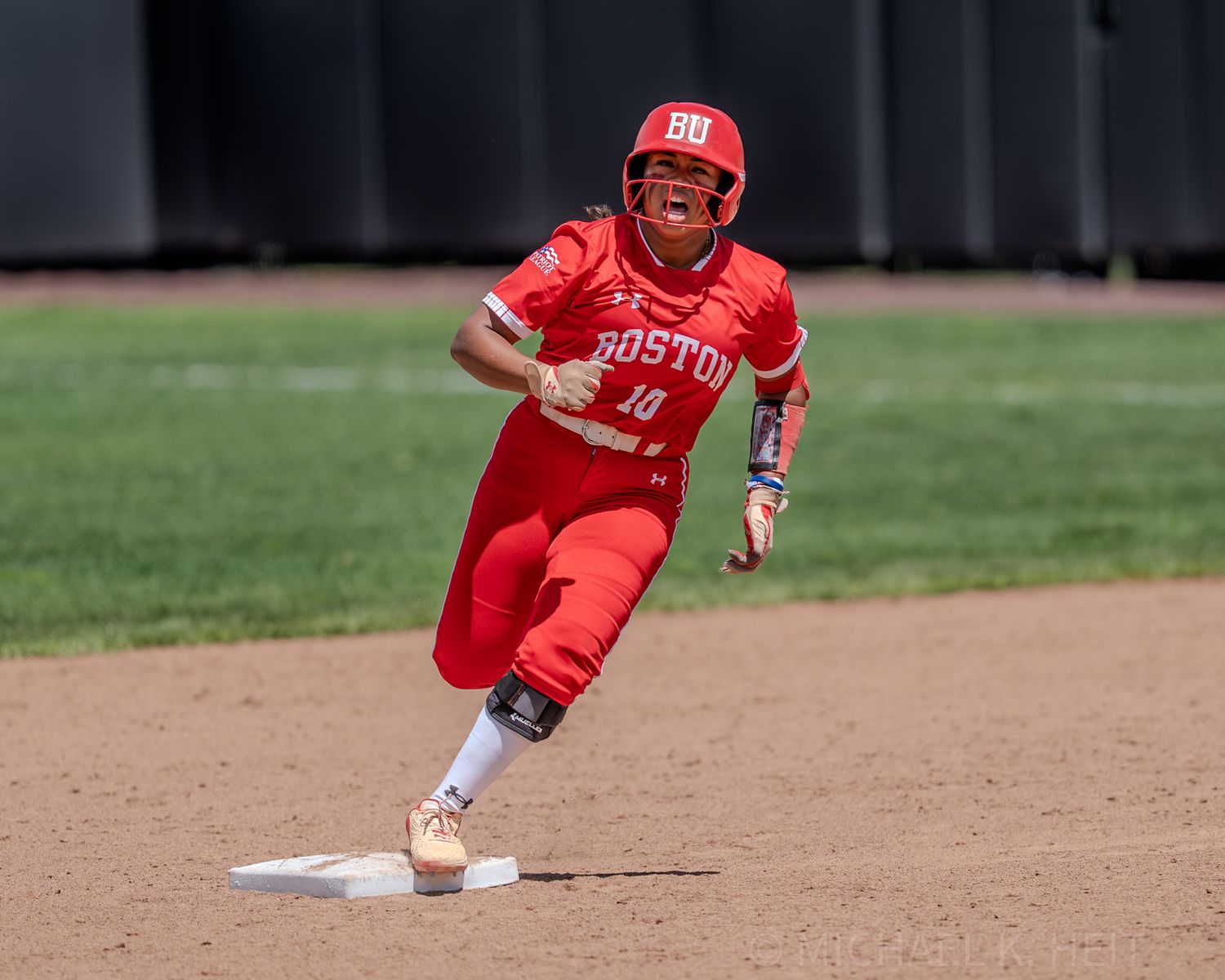 Softball player in red Boston uniform rounds first base during game.