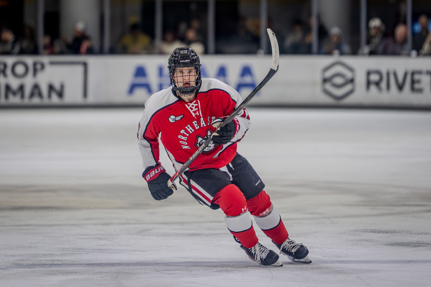 A hockey player in a red jersey and navy blue helmet glides across the ice during a game.