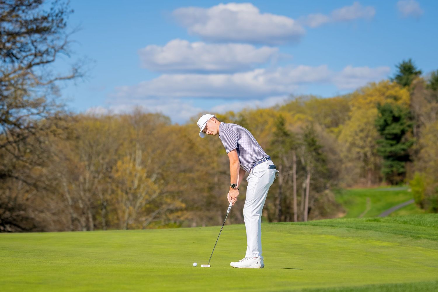 Golfer in white outfit putting on a lush green golf course with trees and blue sky in background.