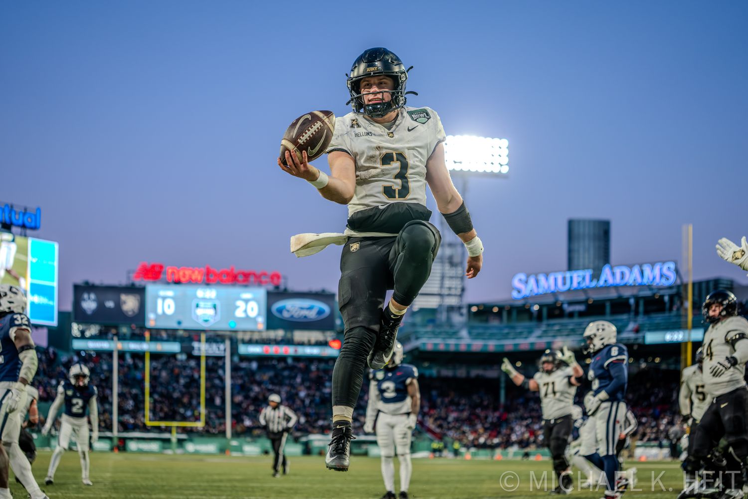 Football players in white and black uniforms compete during a game at a stadium with bright blue sky overhead.