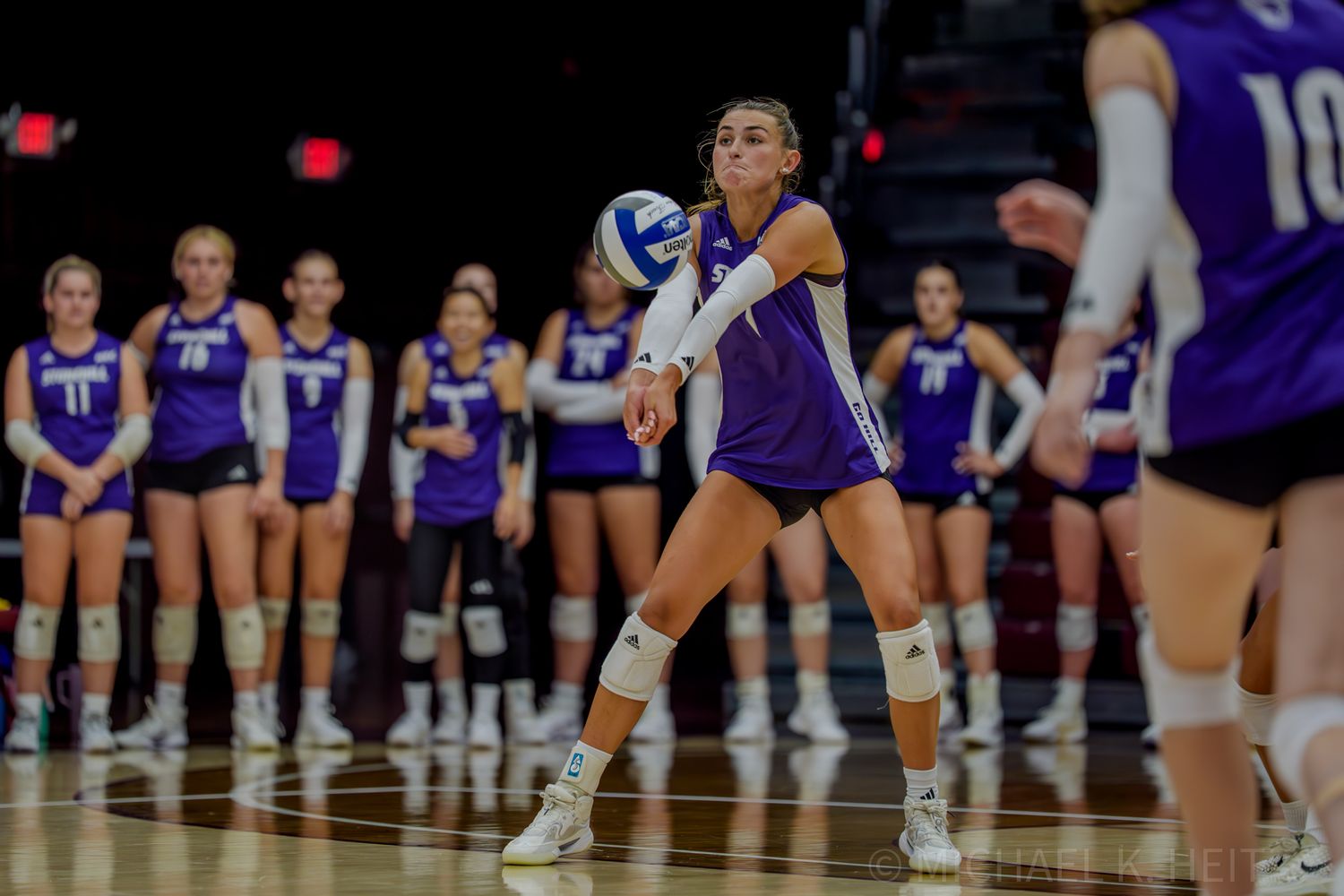 A volleyball player prepares to receive a serve during a match in a purple uniform.