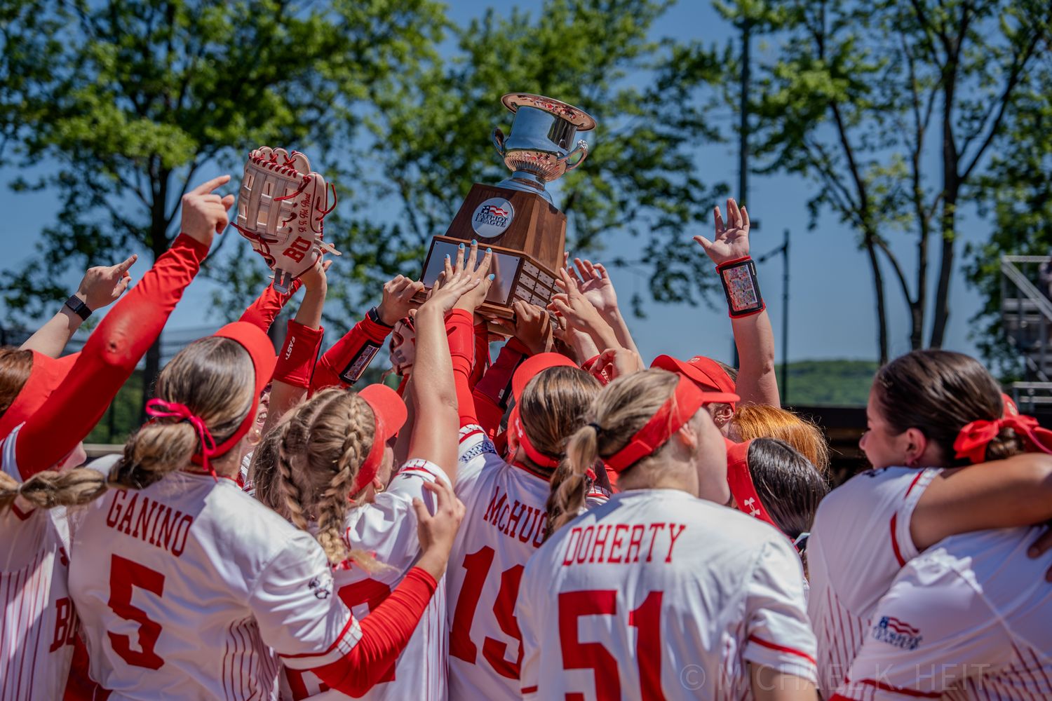 Softball team in red jerseys celebrating with championship trophy overhead.