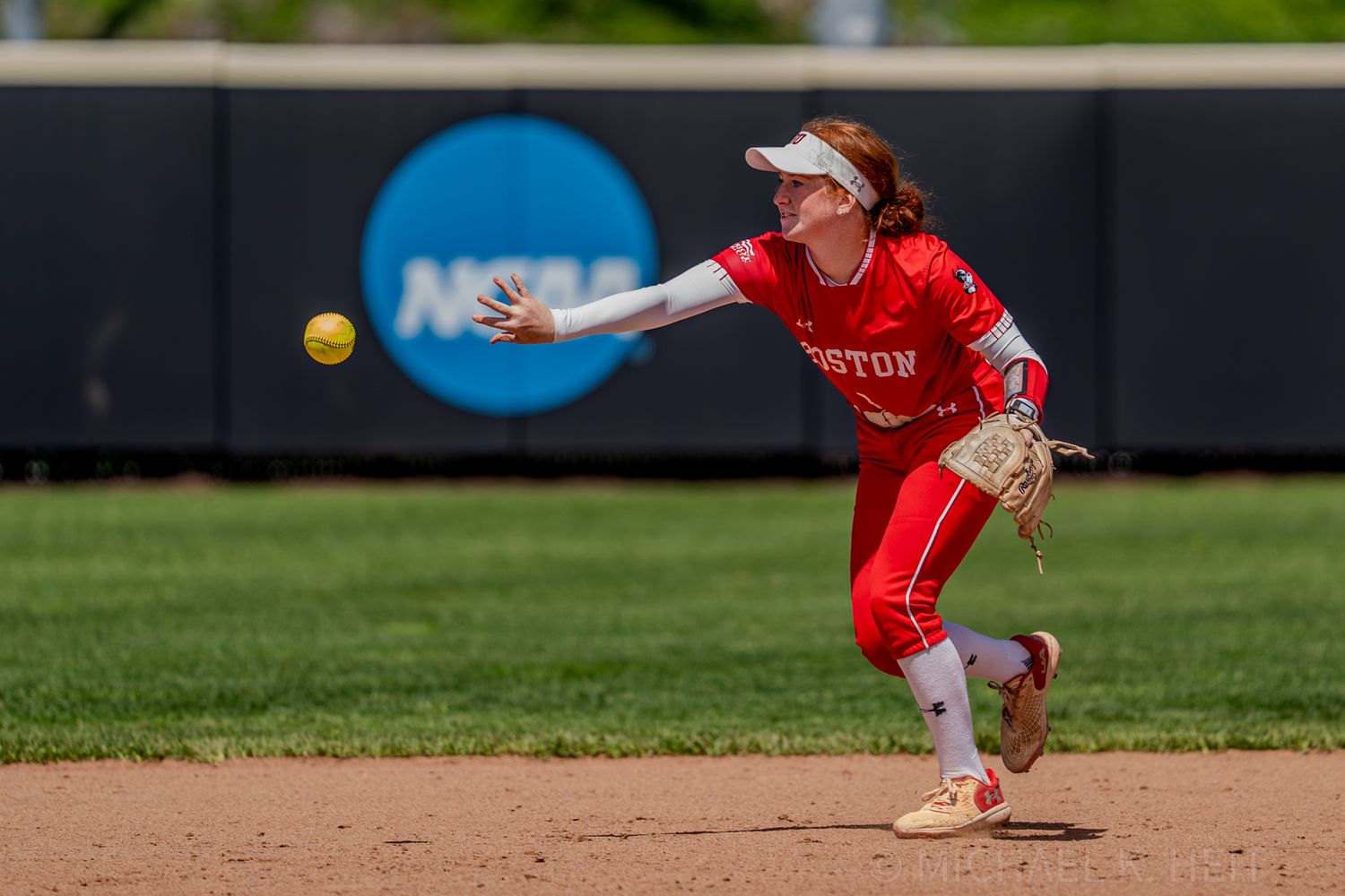 Infielder in red uniform stretches to field ground ball during NCAA softball game.
