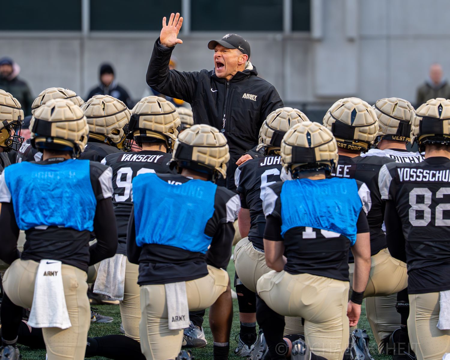 Row of football players in matching uniforms stand on sideline during game.