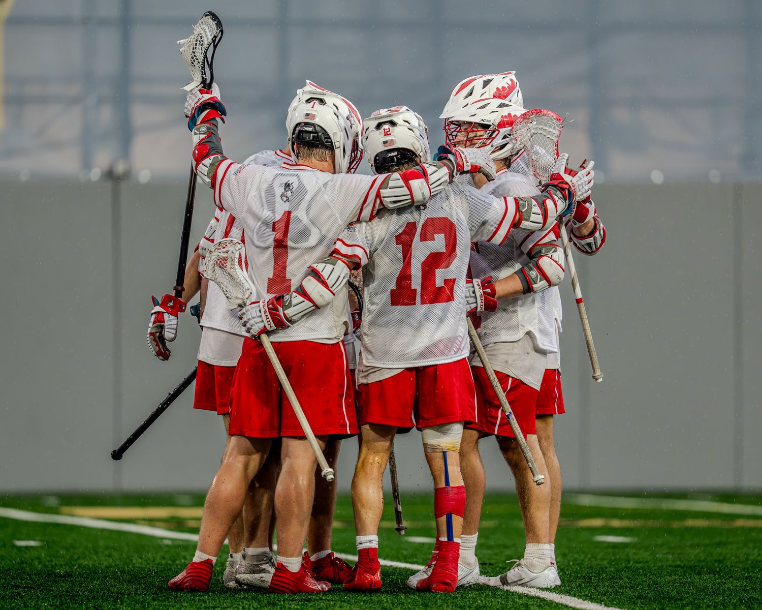 Lacrosse teammates huddle together on the field during a game between teams in white and maroon uniforms.