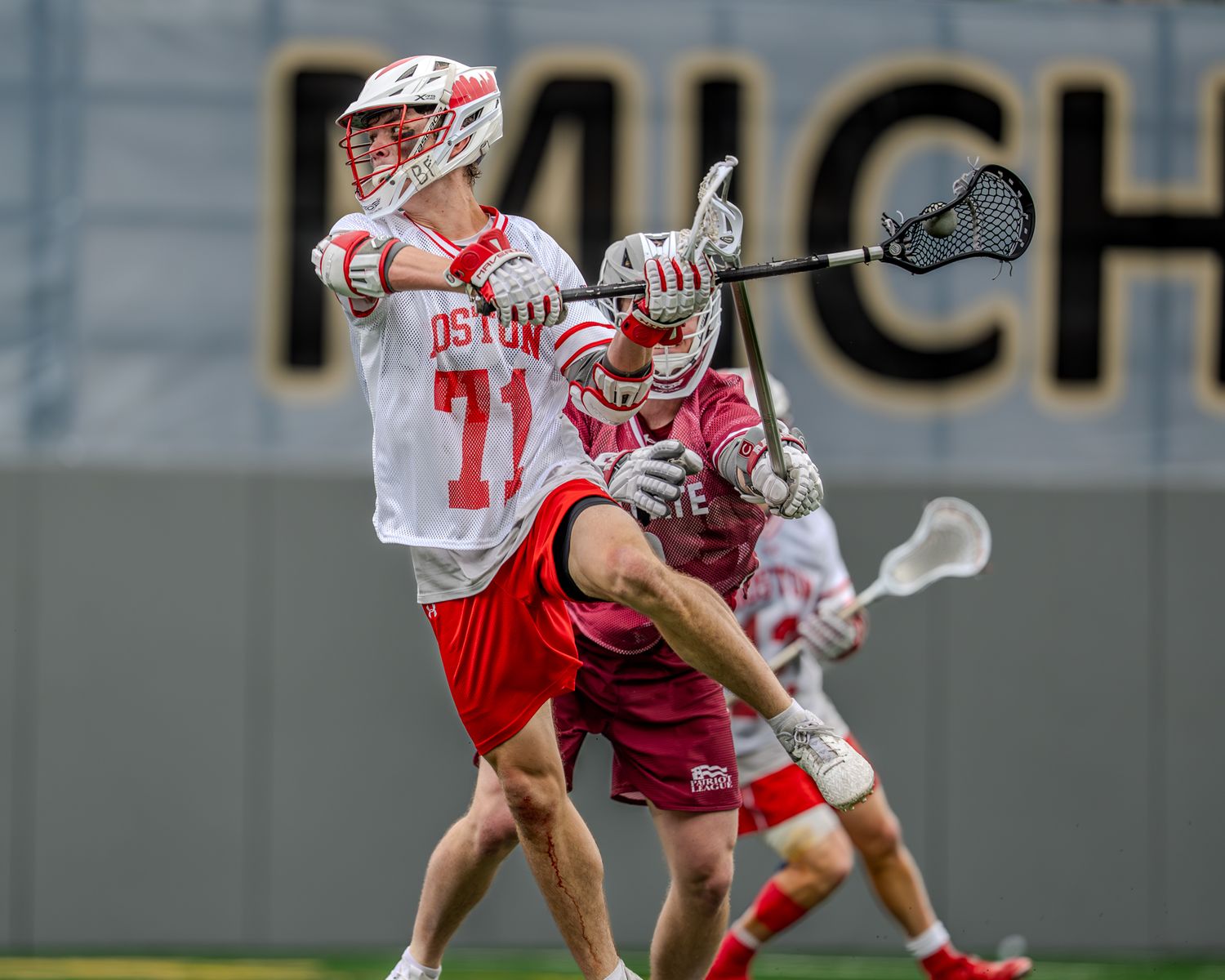 Two collegiate lacrosse teams face off in a competitive match on an artificial turf field.