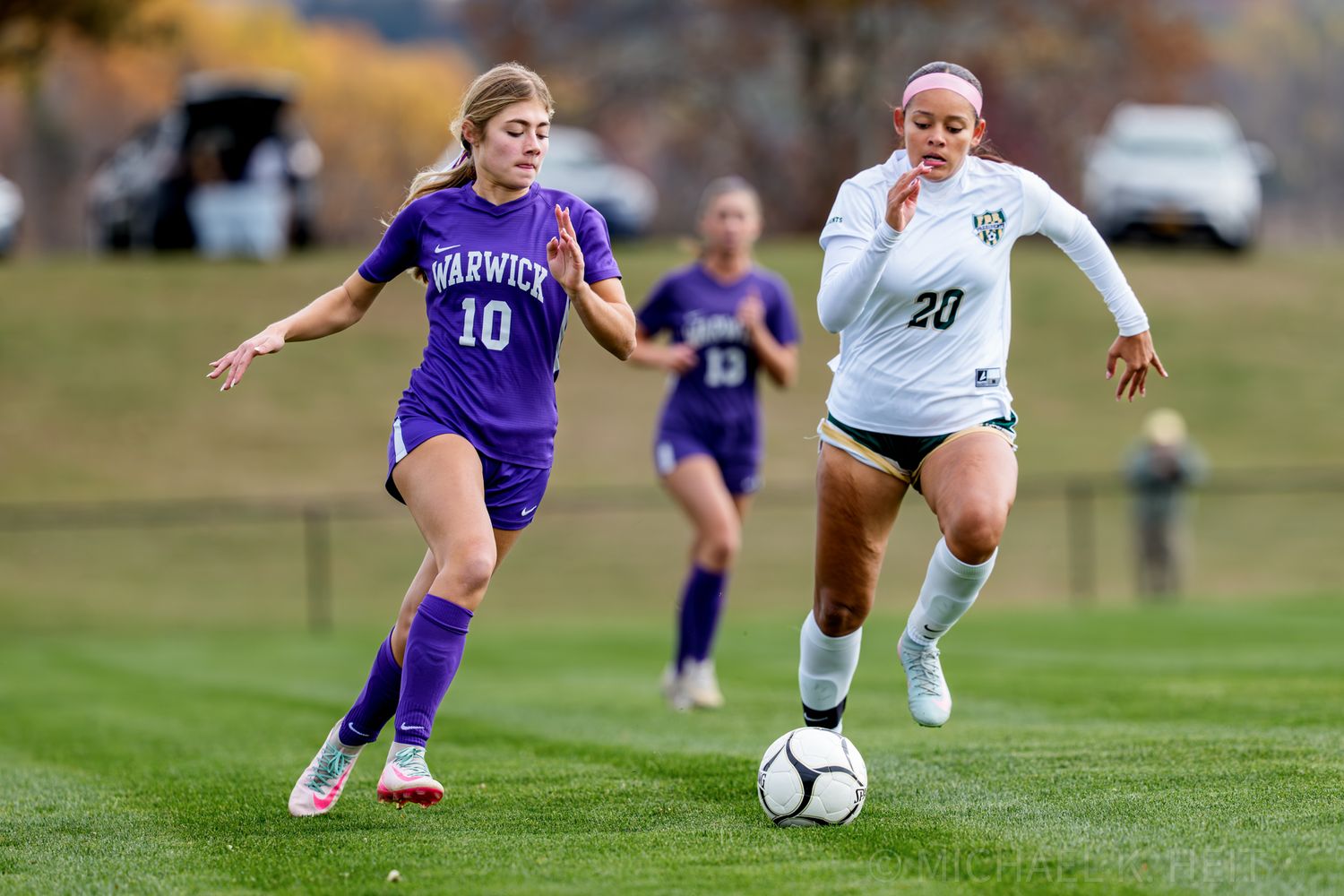 Soccer players compete for possession of the ball during an intense match between teams in white and purple uniforms.