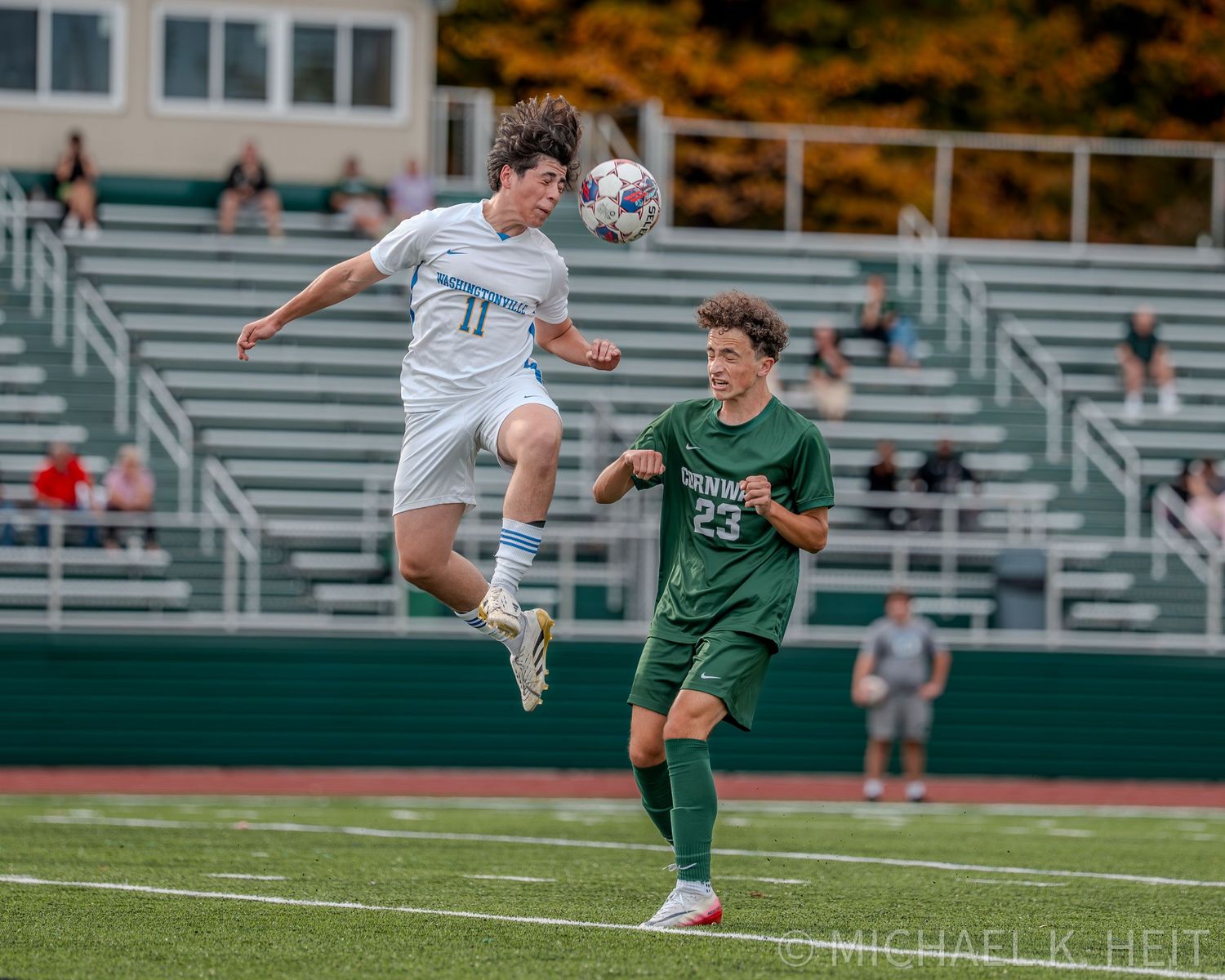 Soccer player in white jersey leaps high to head the ball during competitive match against opponent in green.