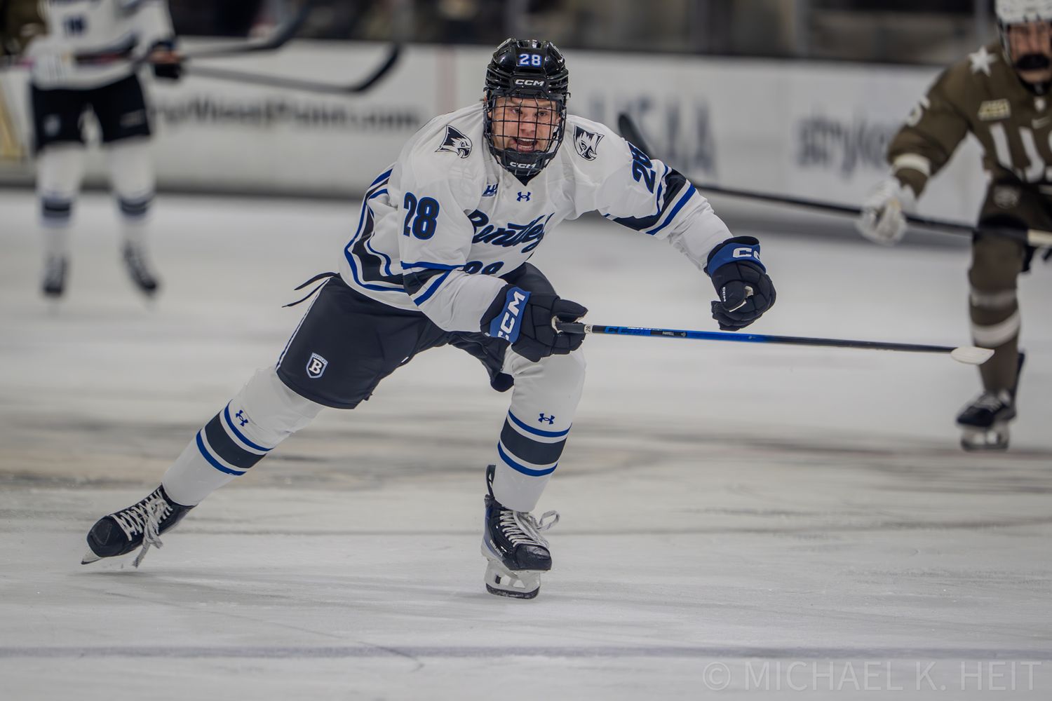 Hockey player in white and blue uniform maintains aggressive skating stance.