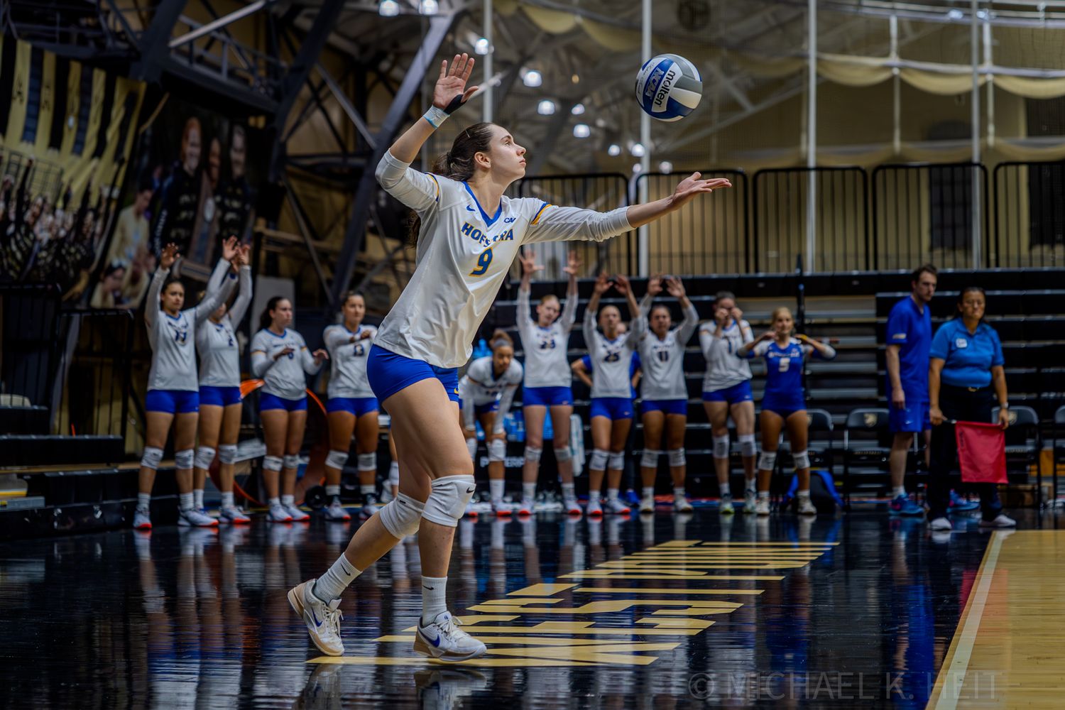 Volleyball players leap at the net during an intense match between two teams in a gymnasium setting.