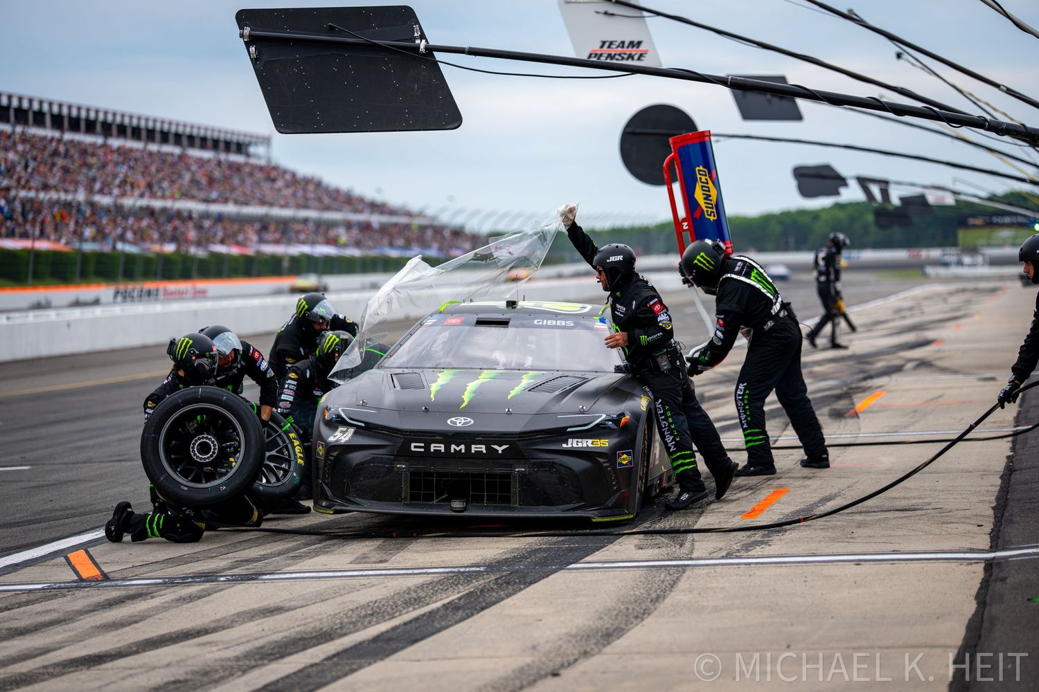 NASCAR pit crew services a black Monster Energy Camry race car during a pit stop at a packed speedway.