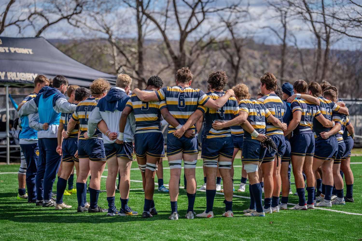 Rugby team huddles together on field during match with coaches providing guidance on sunny day.