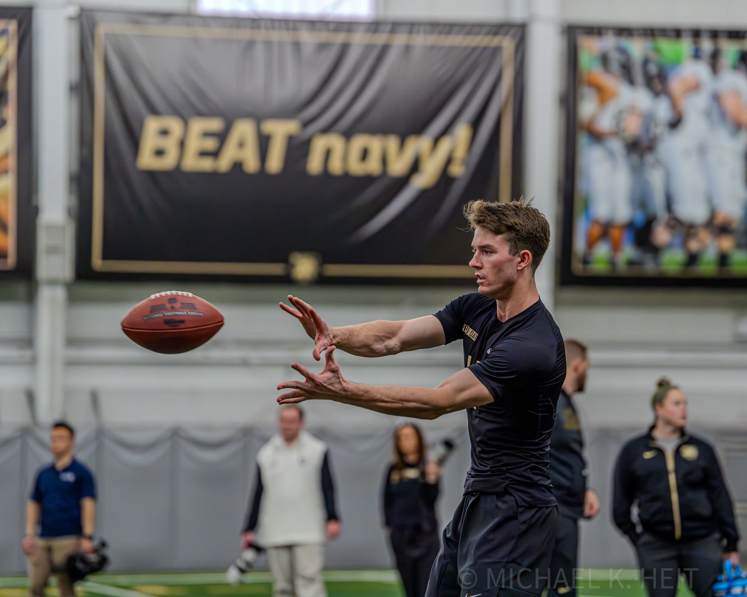 A player practices catching a football in front of a Beat Navy banner.