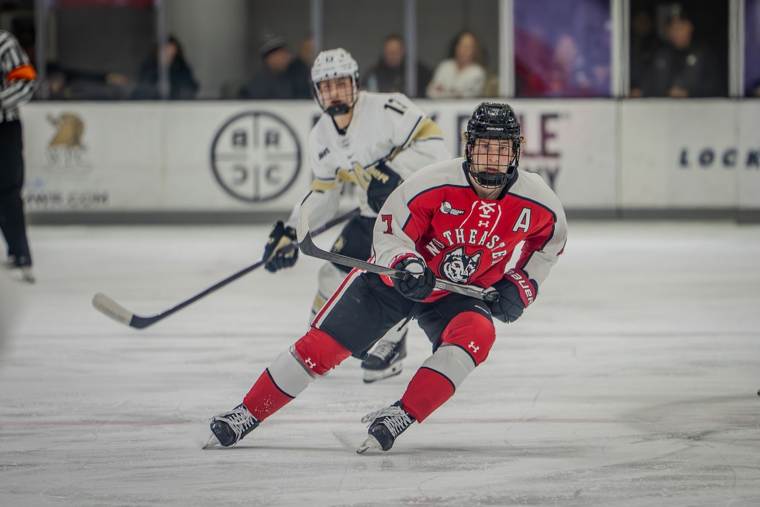 Youth hockey players in red and dark jerseys compete during an ice hockey game at an indoor rink.
