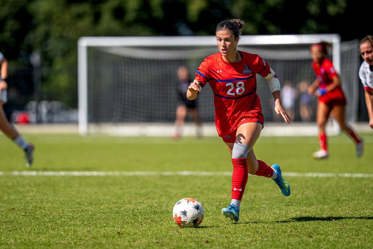 Female soccer player wearing red uniform number 28 dribbling ball on green field during competitive match.