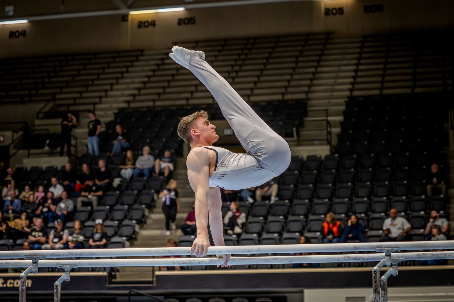 Male gymnast performing a balance skill on parallel bars in a gray uniform at an indoor gymnastics competition.
