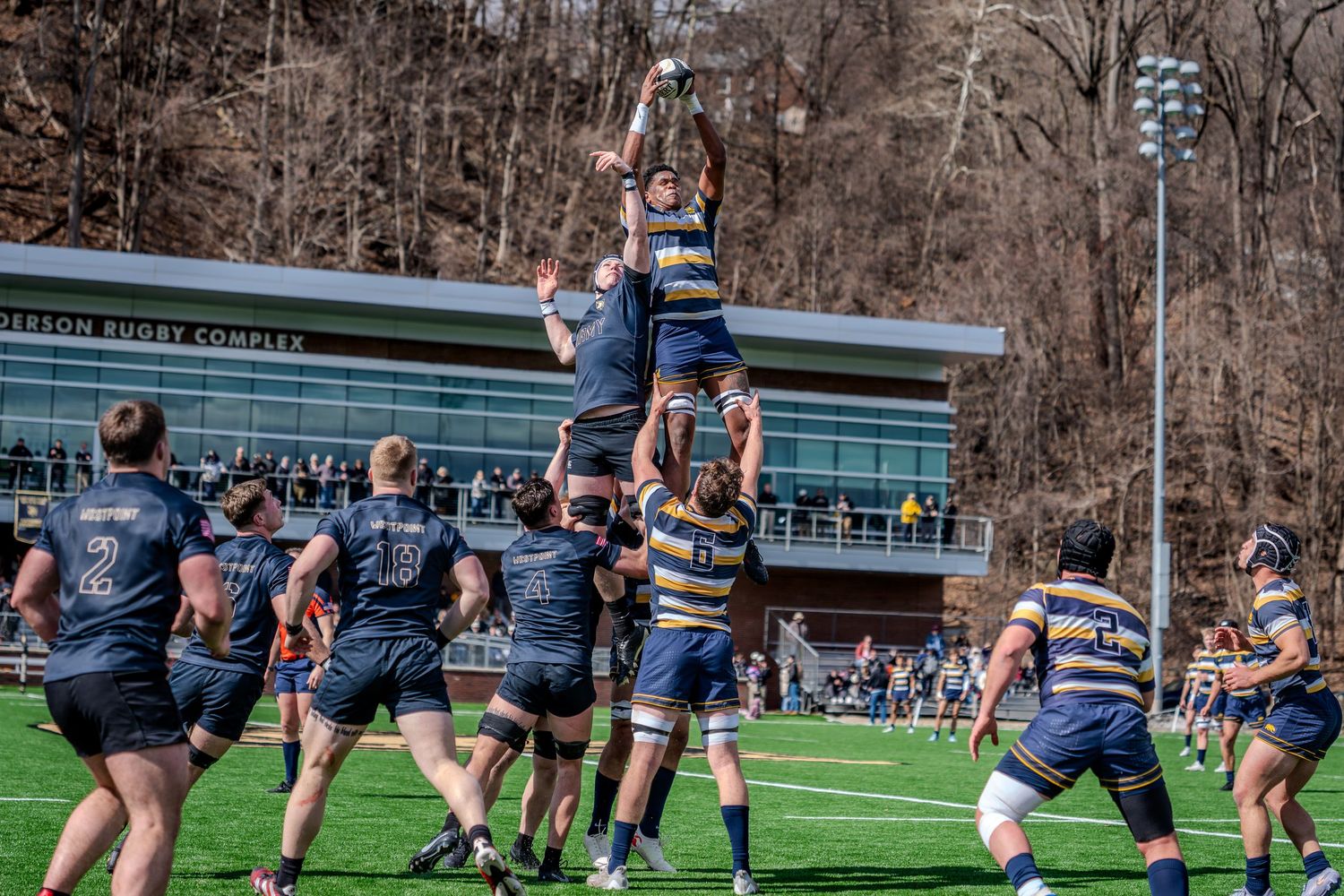 Rugby players compete for possession during an intense match on a turf field with spectators watching from sidelines.