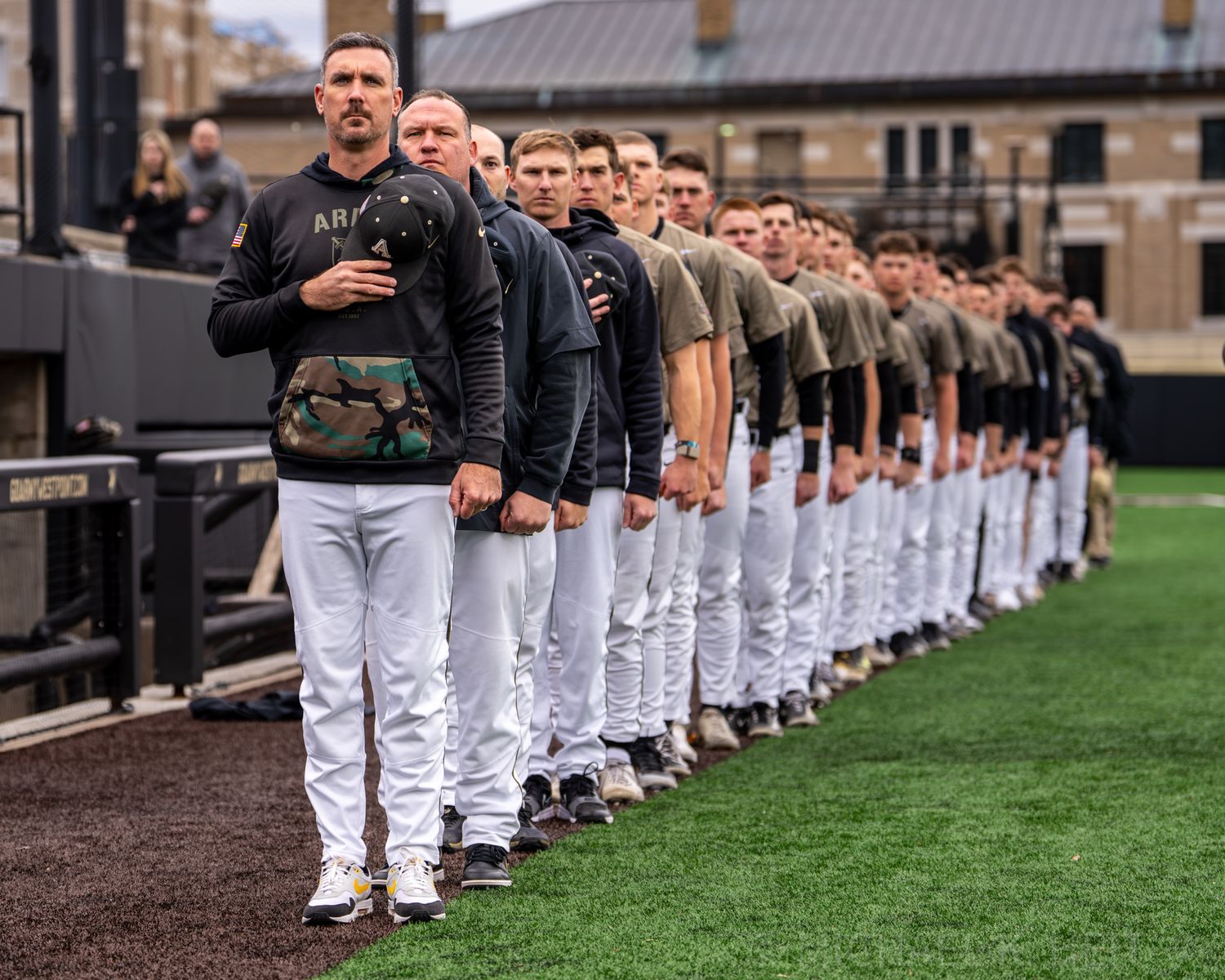 Baseball team members line up in white uniforms along the field sideline.