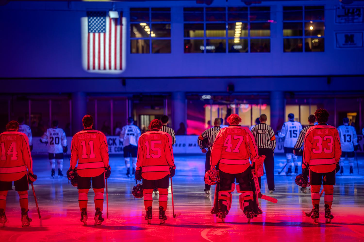 Hockey players stand in line on ice rink during national anthem ceremony with American flag displayed on screens.