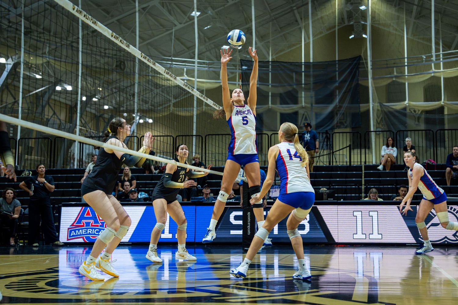 College volleyball player in blue jersey sets the ball during a competitive indoor match with teammates and opponents nearby.