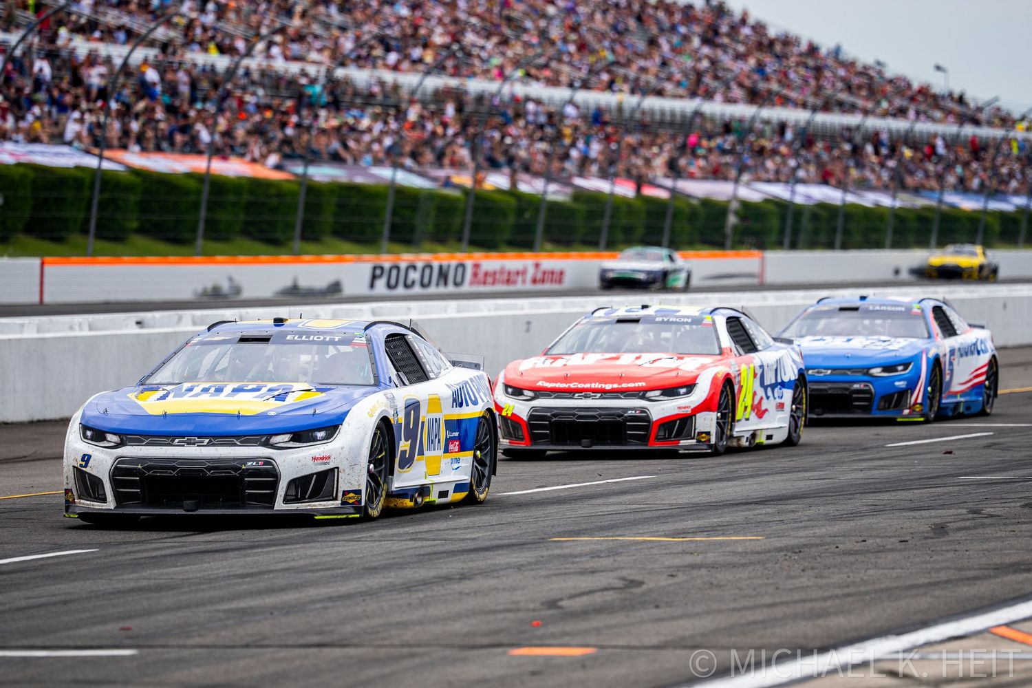 NASCAR race cars compete on the Pocono Raceway track with packed grandstands of fans watching the high-speed action.