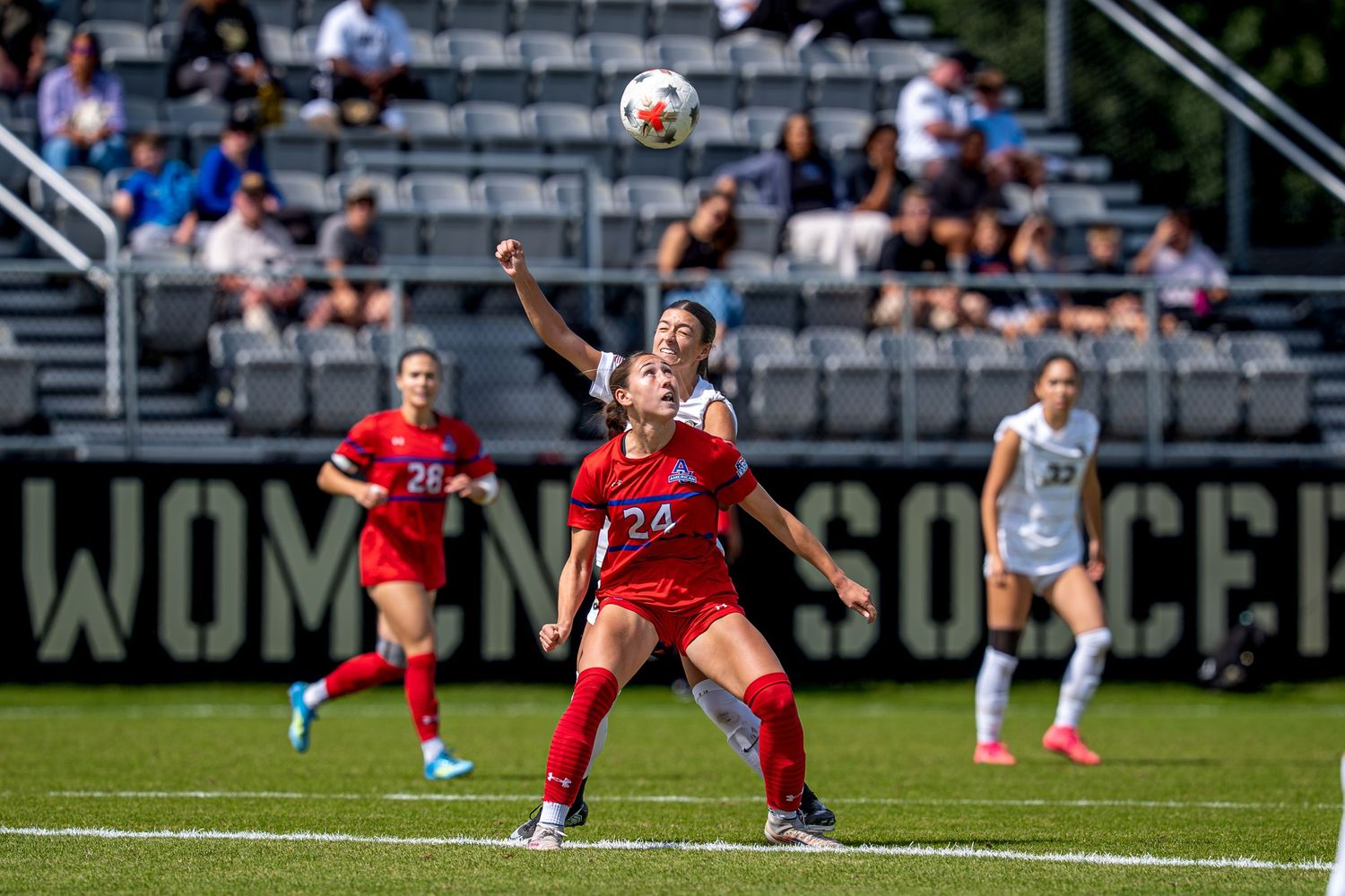 Female soccer players compete for aerial ball during a women's college soccer match on a sunny day.