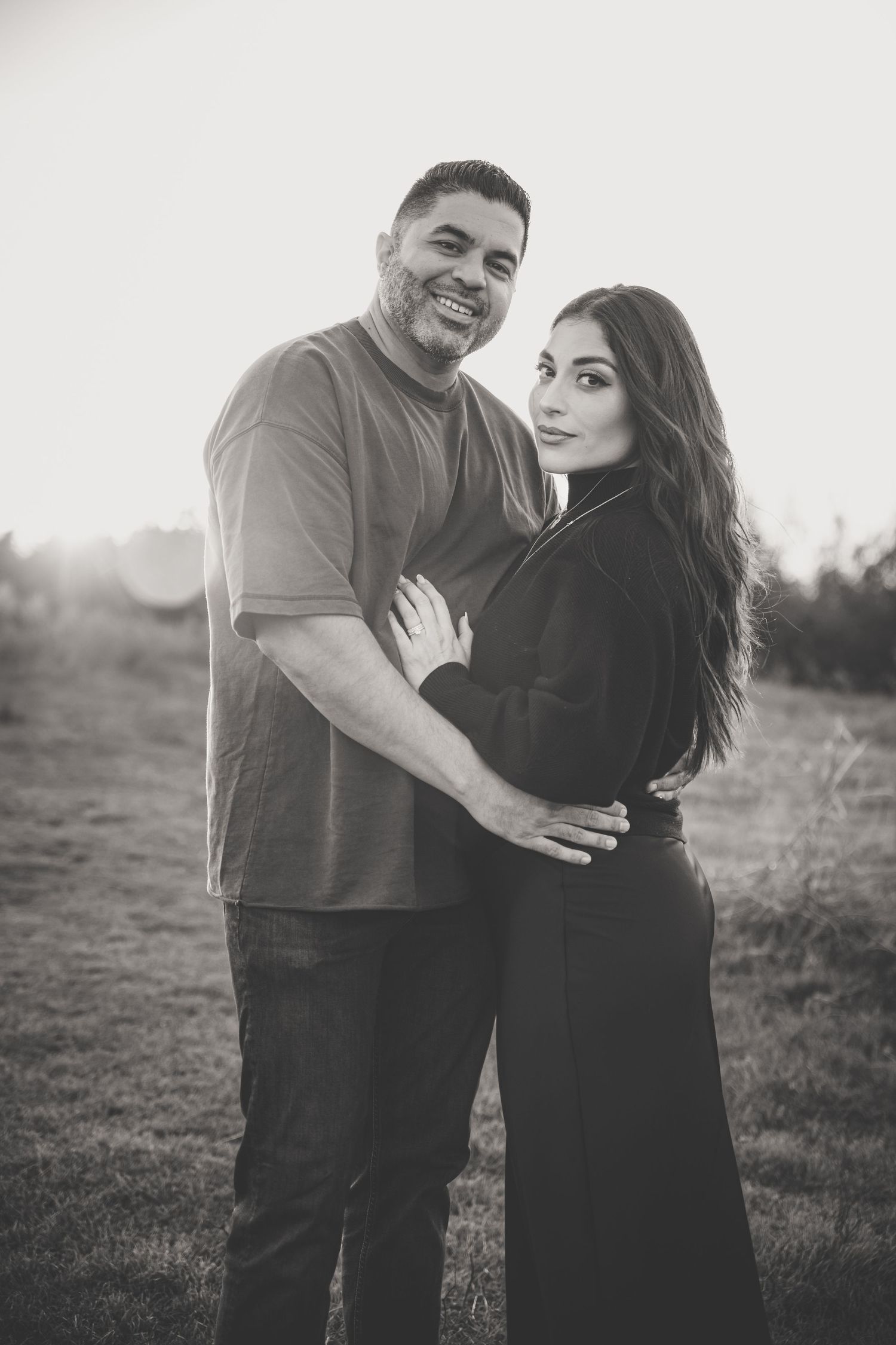 Black and white portrait of a smiling couple embracing in a sunlit outdoor field at golden hour.