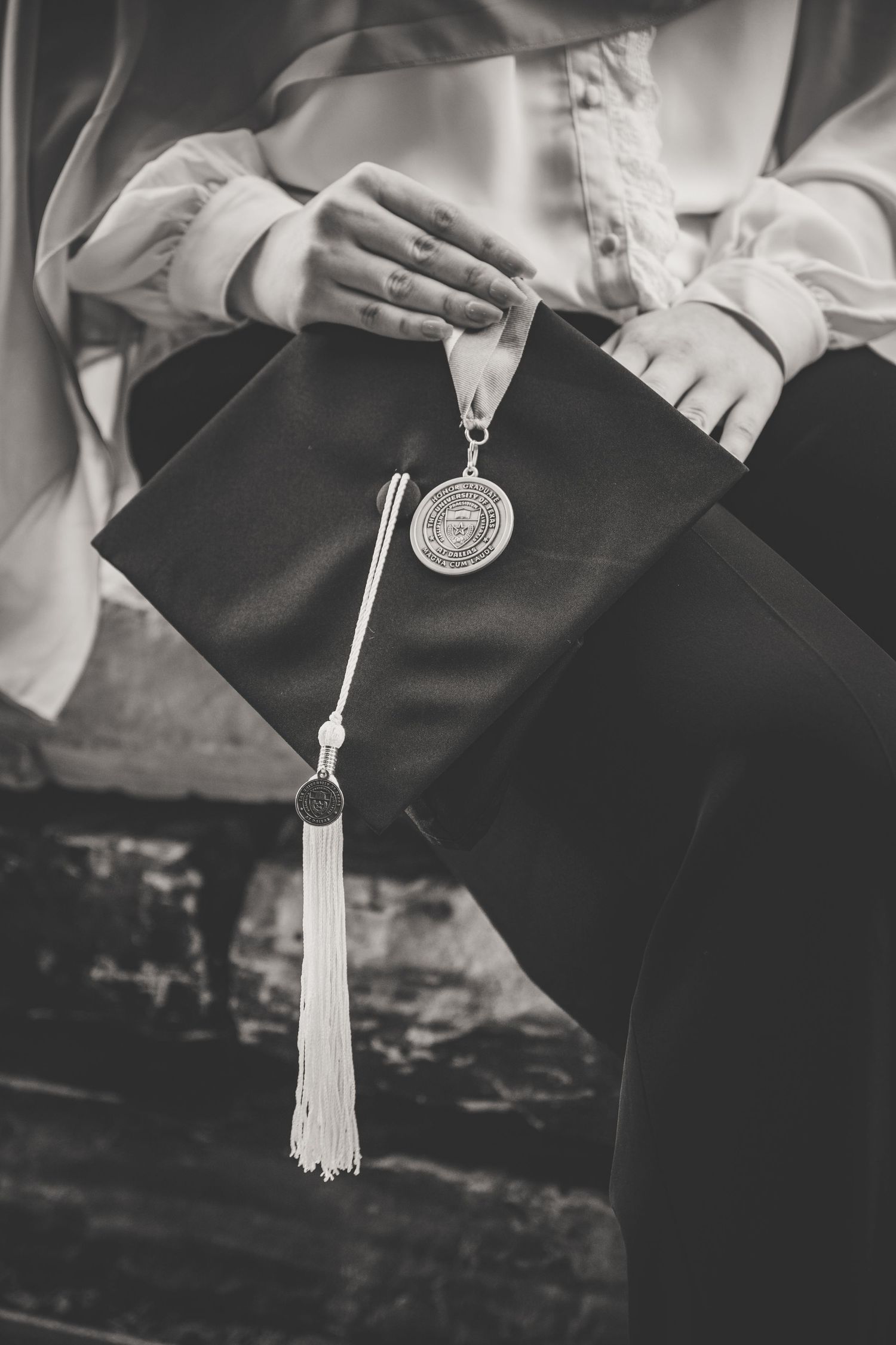 A black and white photo of hands holding a graduation cap with a medal and tassel.
