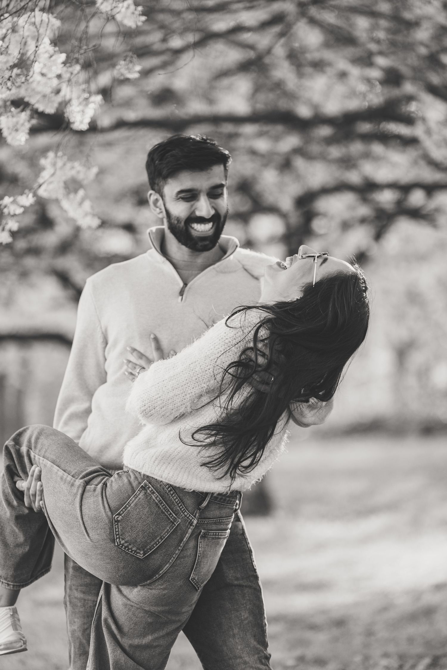 Black and white photo of a man joyfully lifting a laughing woman under blooming cherry blossom trees.