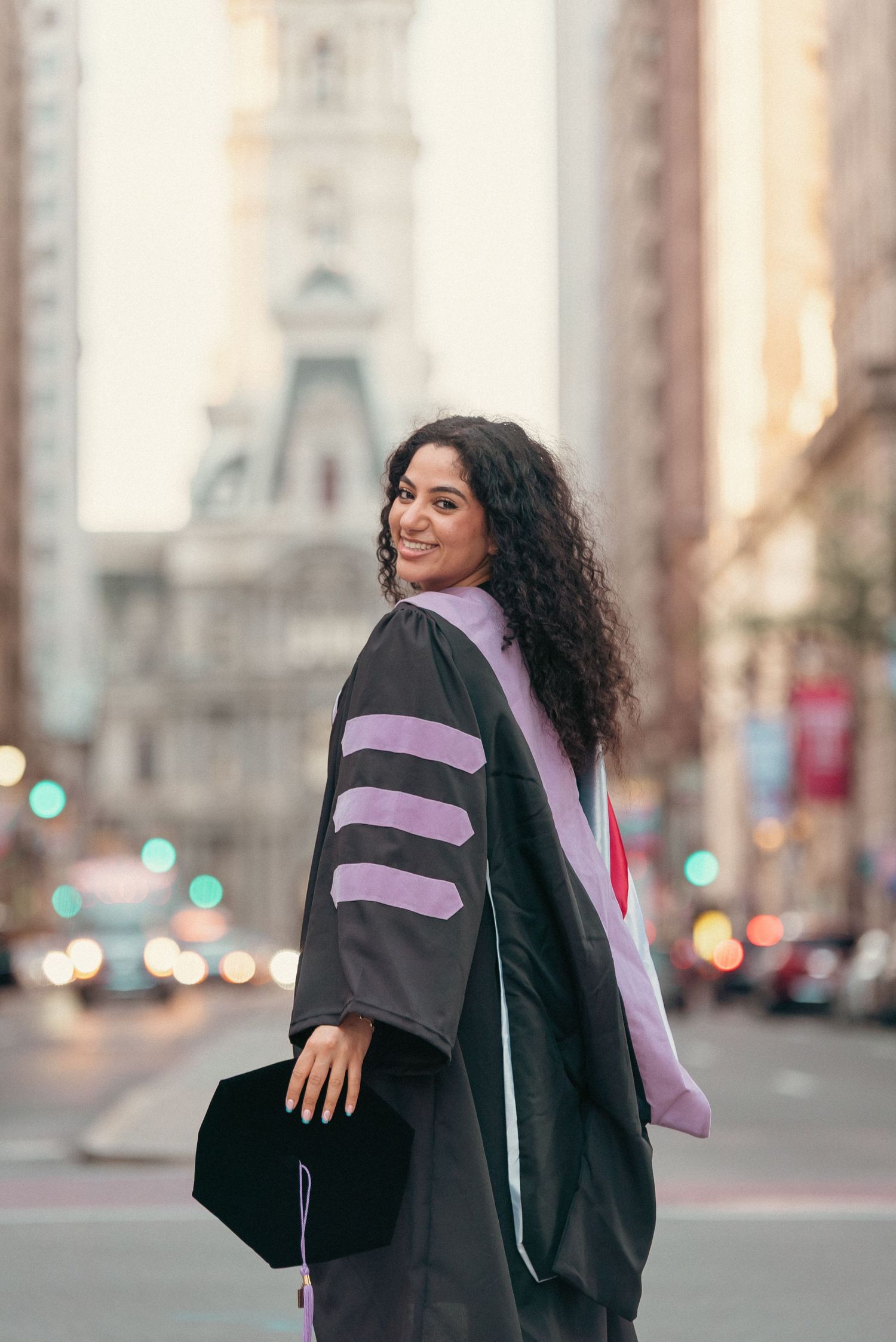 Graduate in doctoral regalia smiling over her shoulder on a city street with urban buildings in the background.