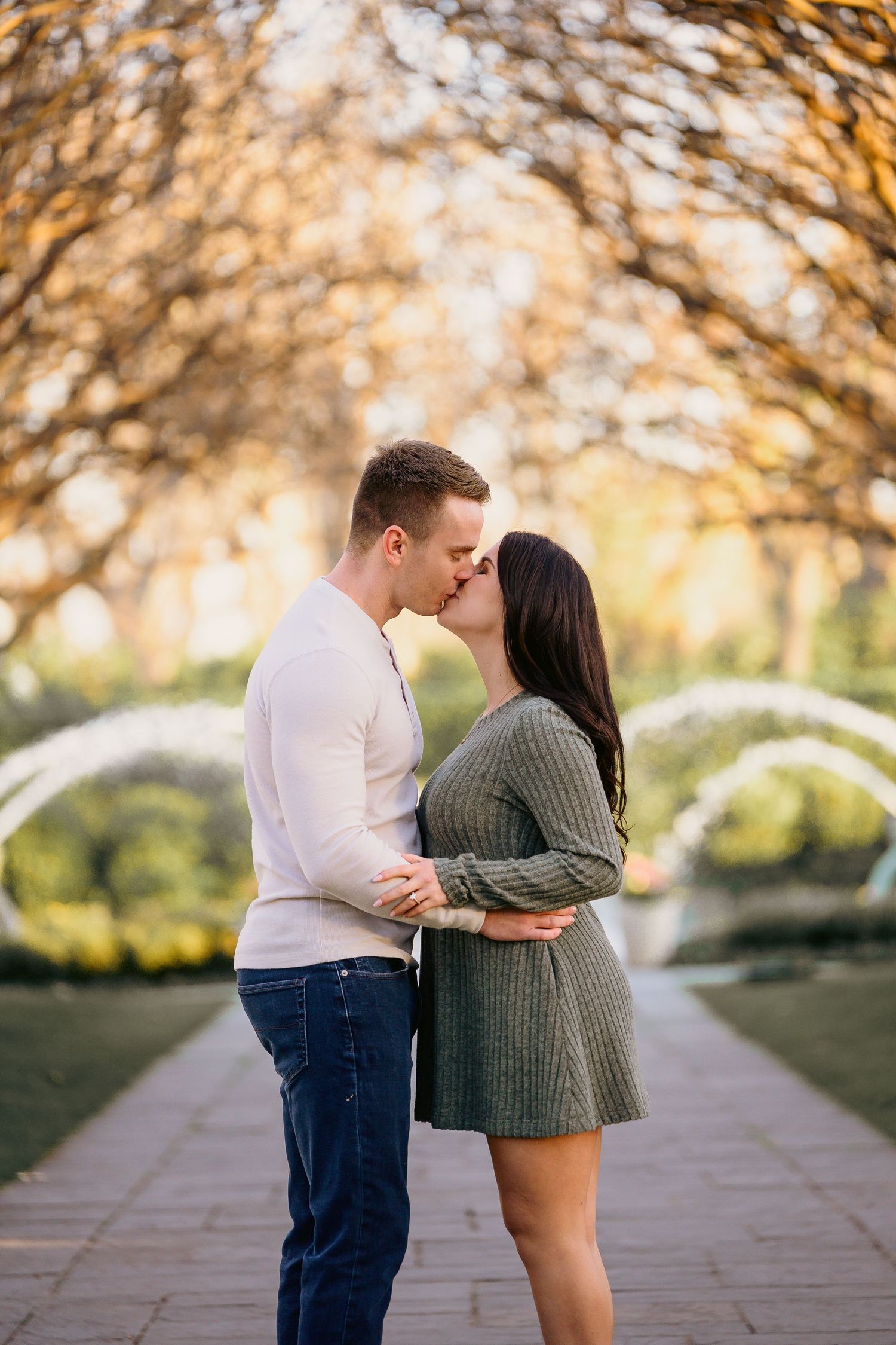 A couple shares a romantic kiss on a tree-lined pathway in a golden autumn park.