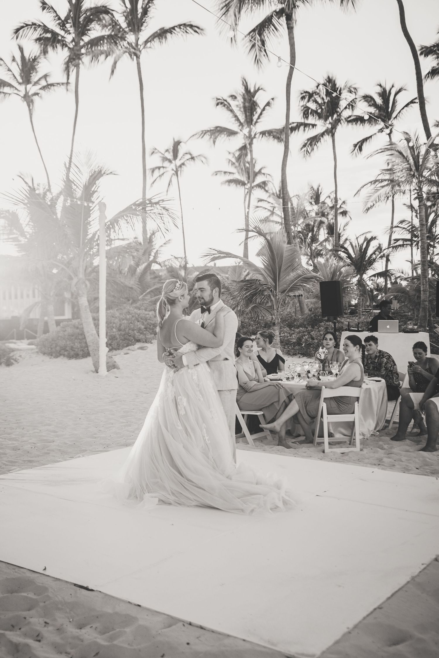 Bride and groom share first dance on sandy beach surrounded by palm trees and wedding guests.