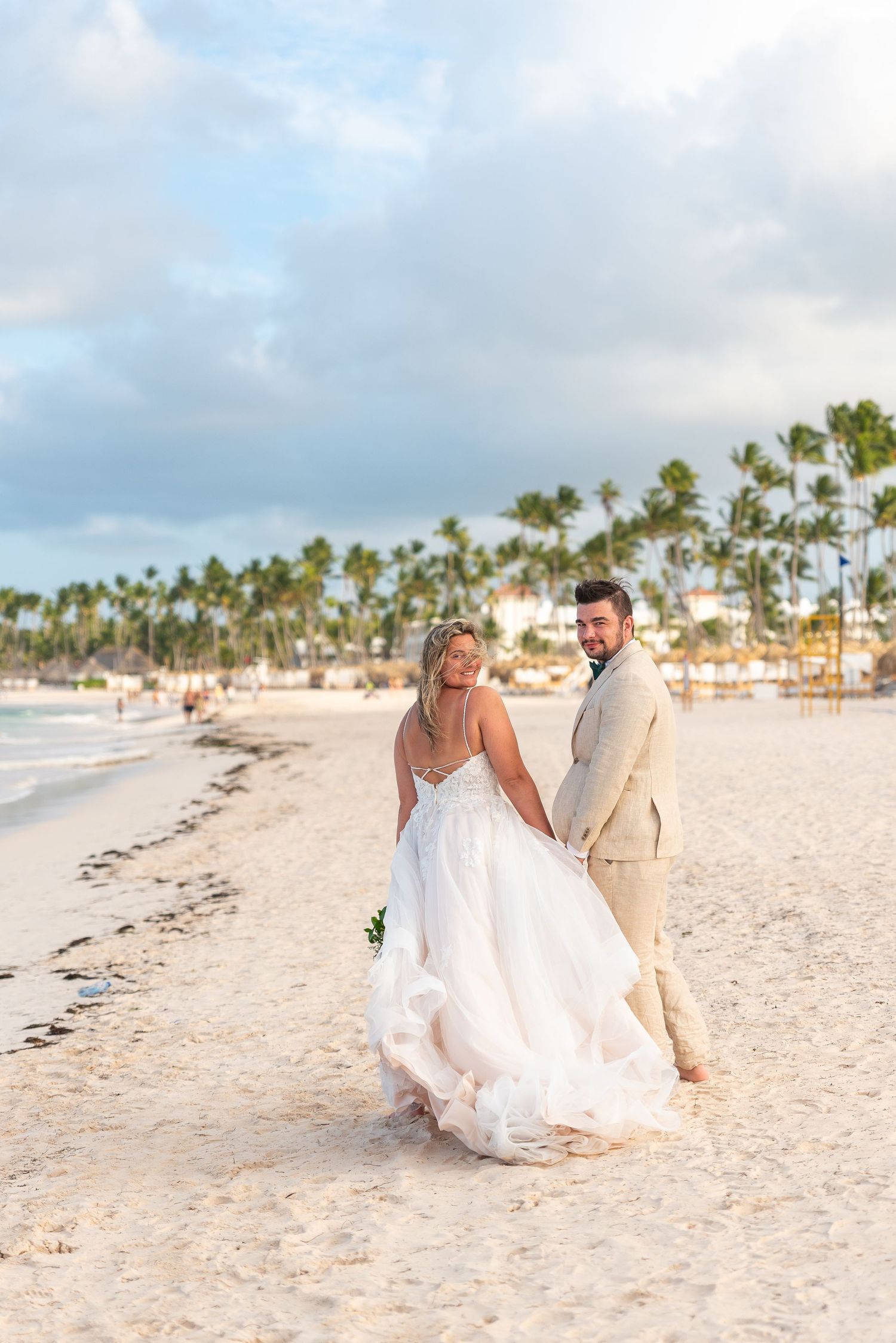 Bride and groom walk hand in hand along white sand beach with palm trees at sunset.