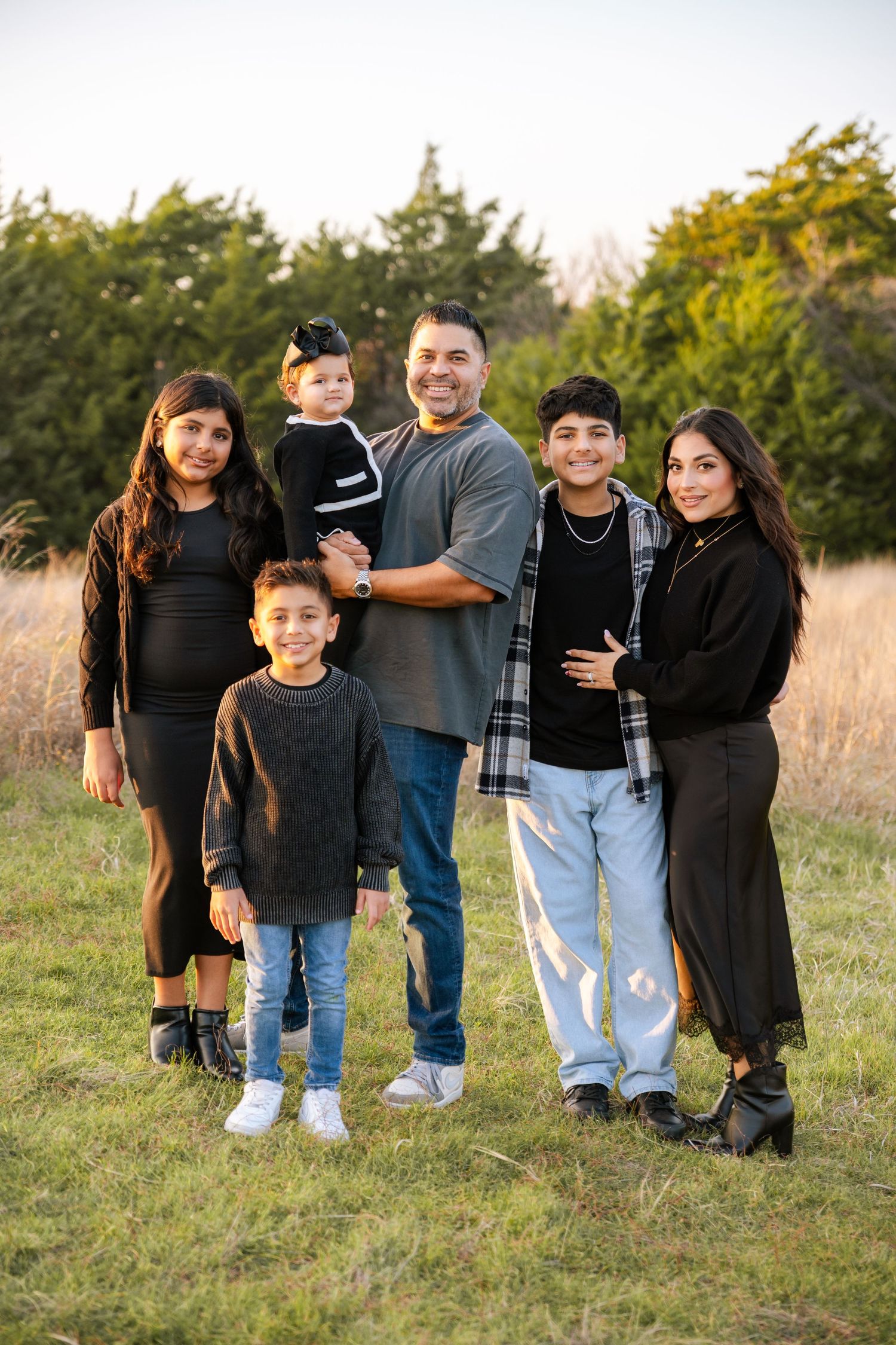 A joyful family of six poses together in a golden field with evergreen trees behind.