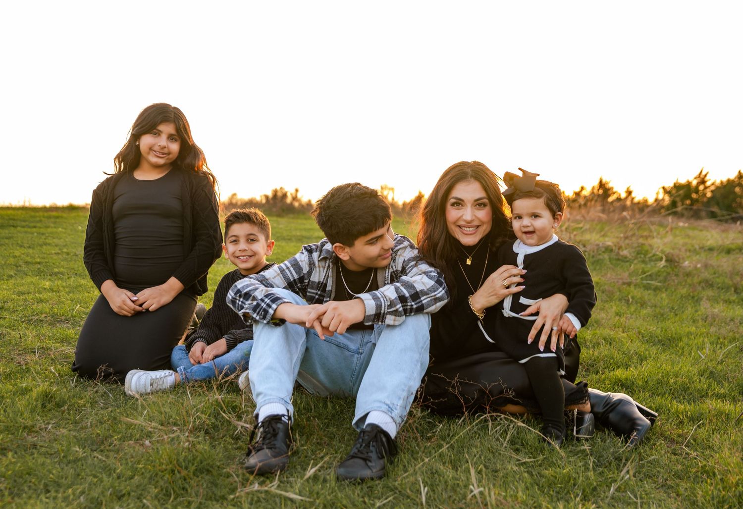 Family of five sitting together in a golden field at sunset, smiling warmly in coordinated black outfits.
