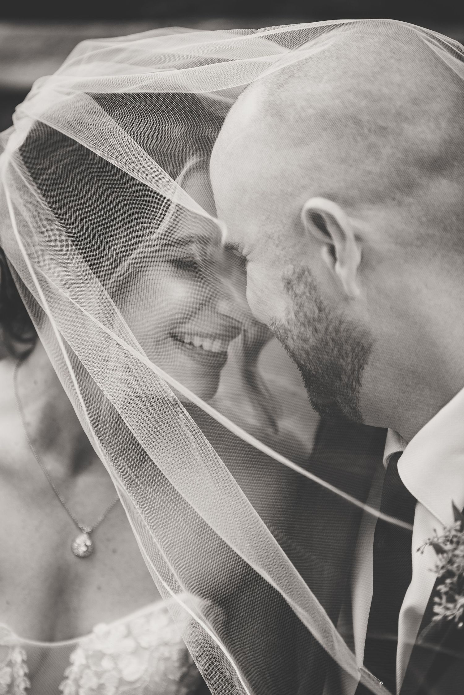 Black and white wedding photo of smiling bride and groom sharing an intimate moment under a flowing veil.