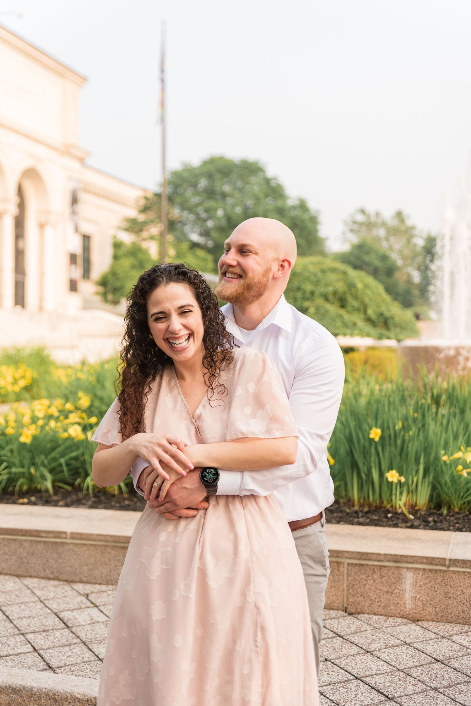Happy couple embracing outdoors near yellow flowers, woman in pink dress, man in white shirt.