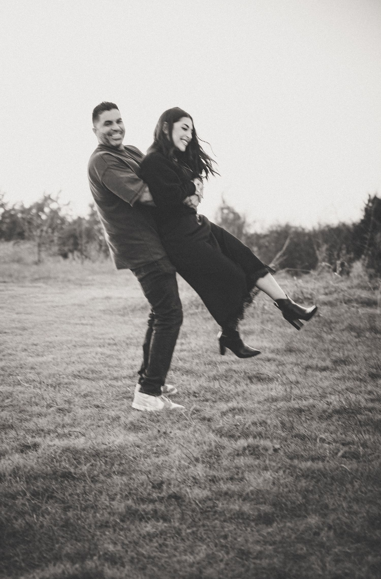 A playful black and white photo of a man lifting and spinning a laughing woman in an open field.