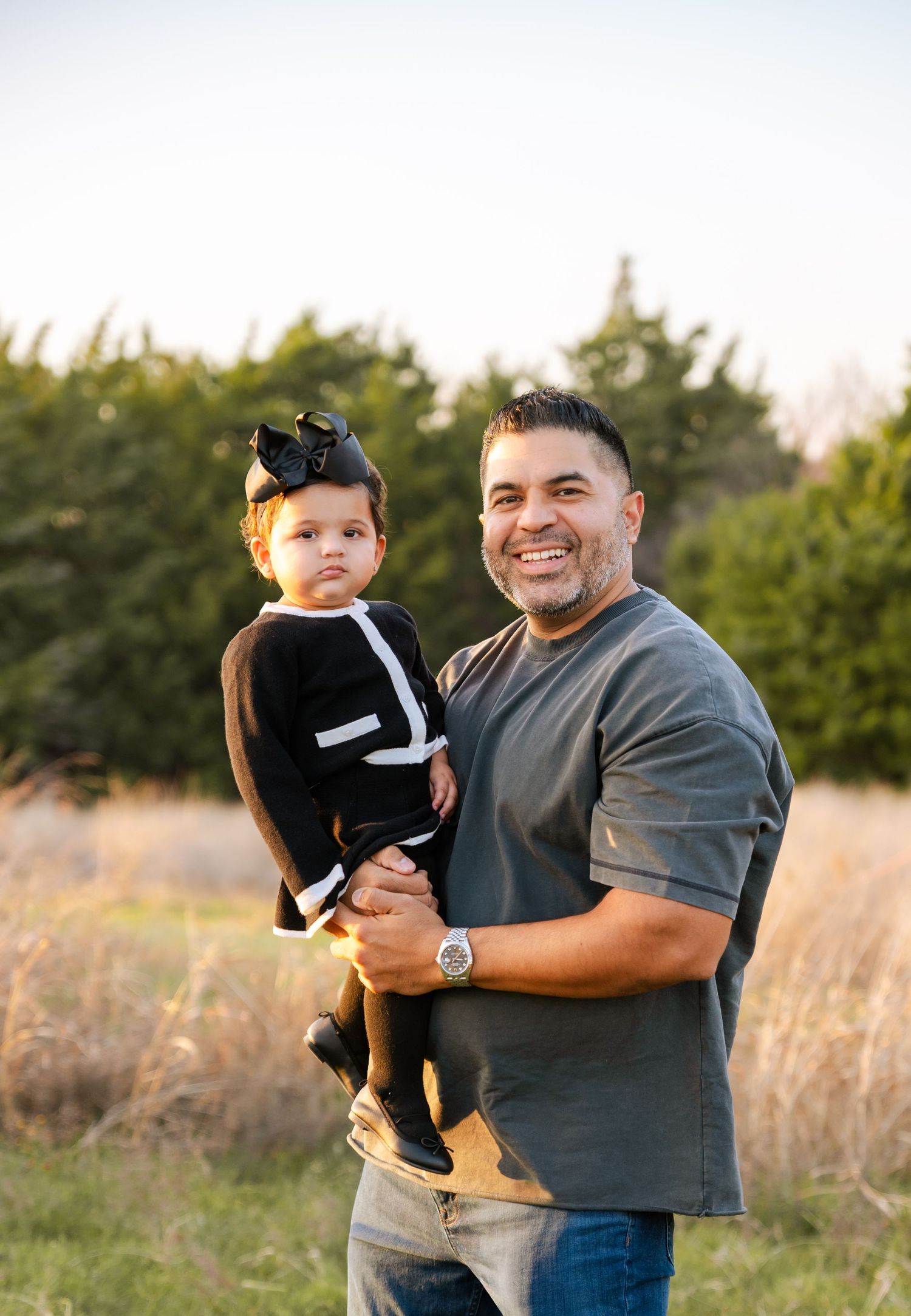 A smiling father holds his baby daughter in a sunlit field near evergreen trees.