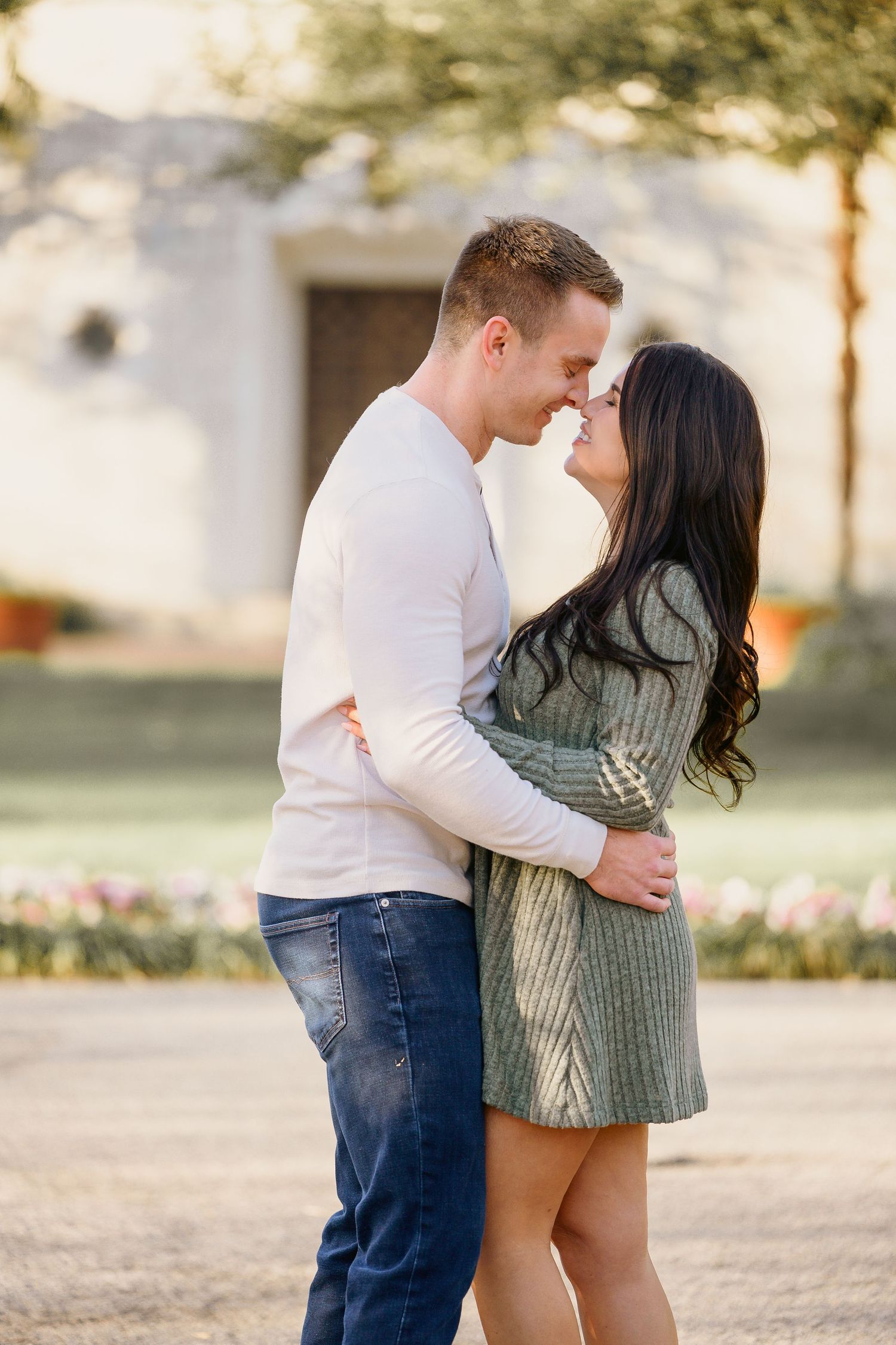 A couple embraces and nearly kisses outdoors in warm golden sunlight with flowers in the background.