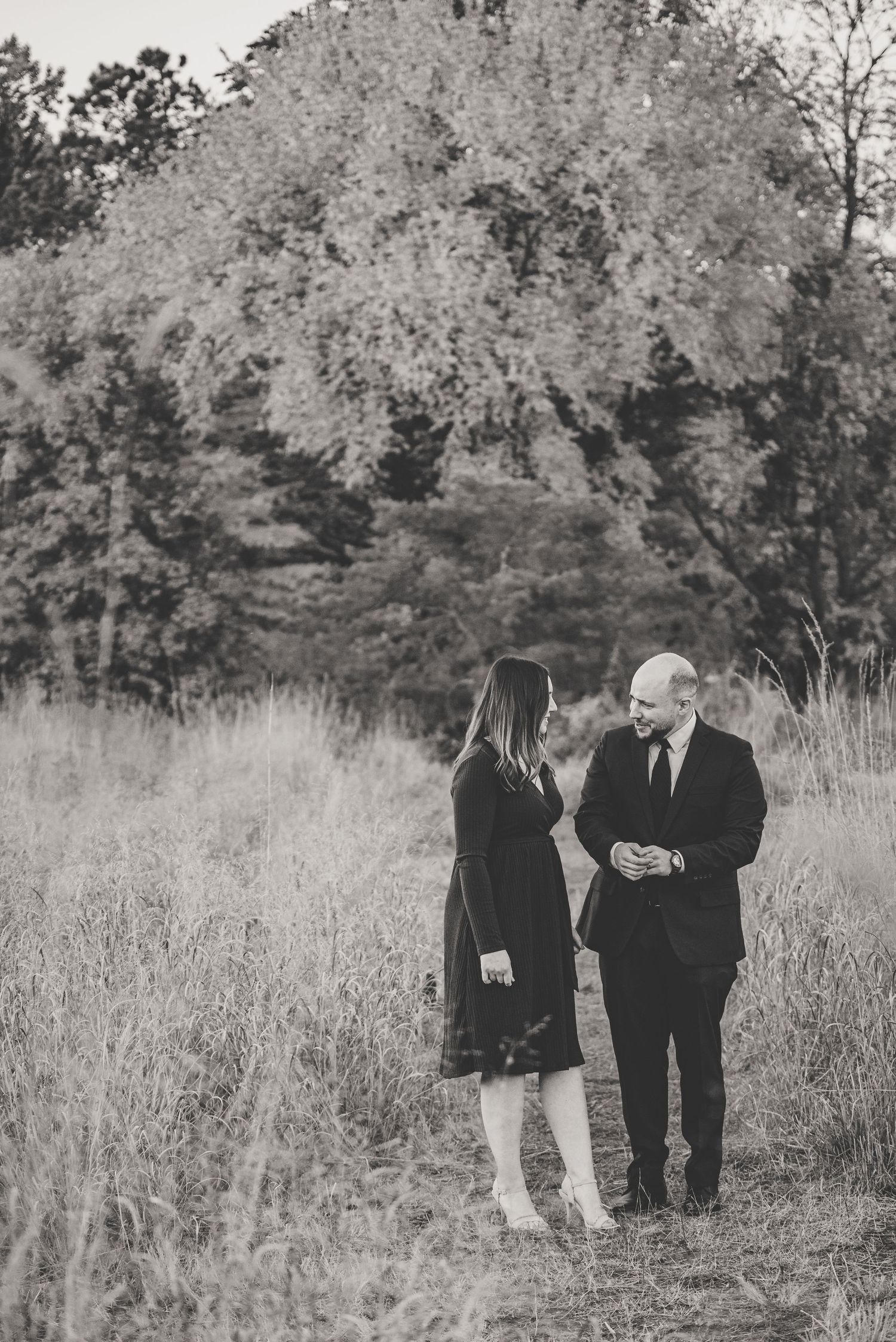 Black and white photo of a couple standing in a field of tall grass, sharing a romantic moment with trees in the background.