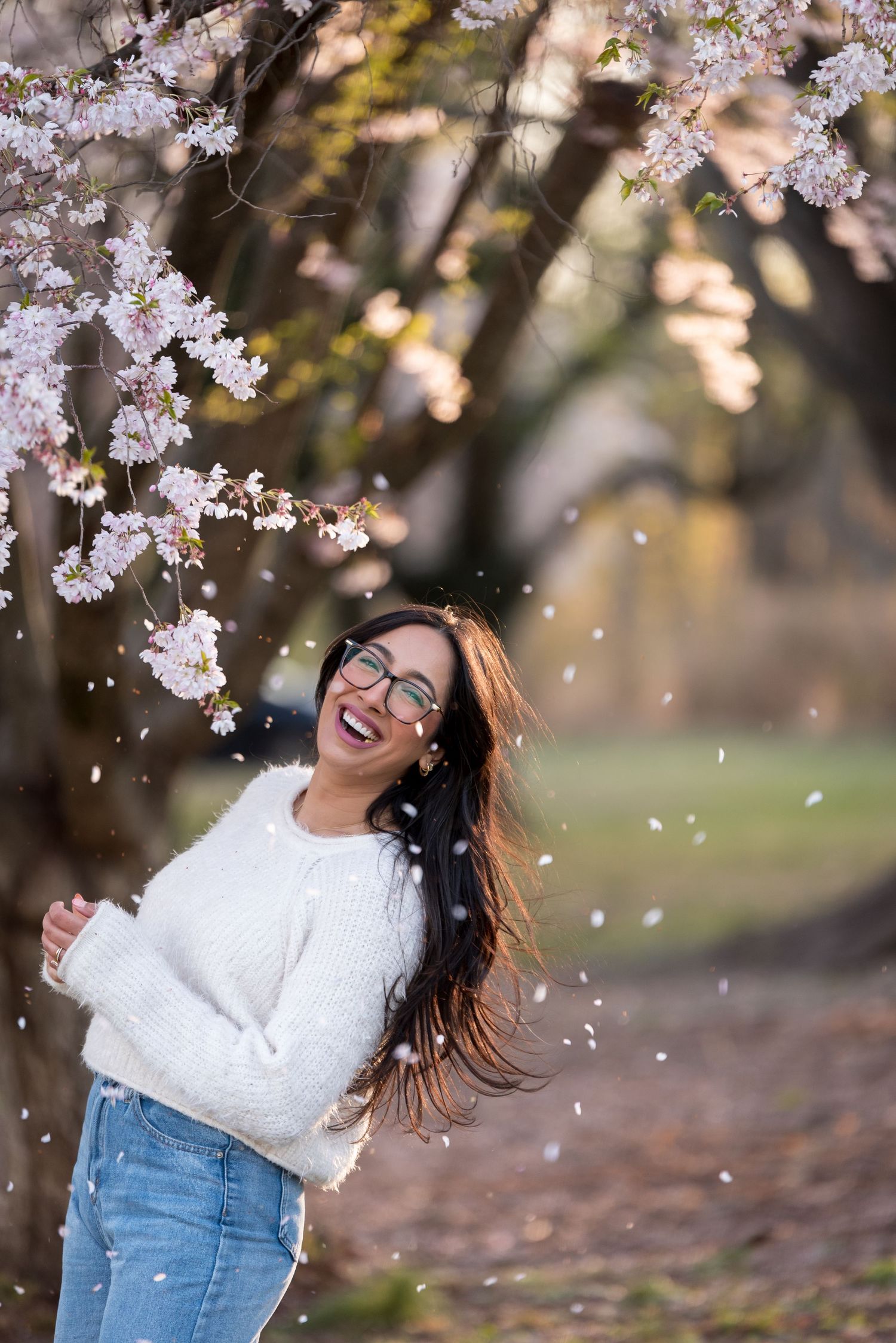 A joyful woman laughs under blooming cherry blossom trees as petals fall around her in spring.