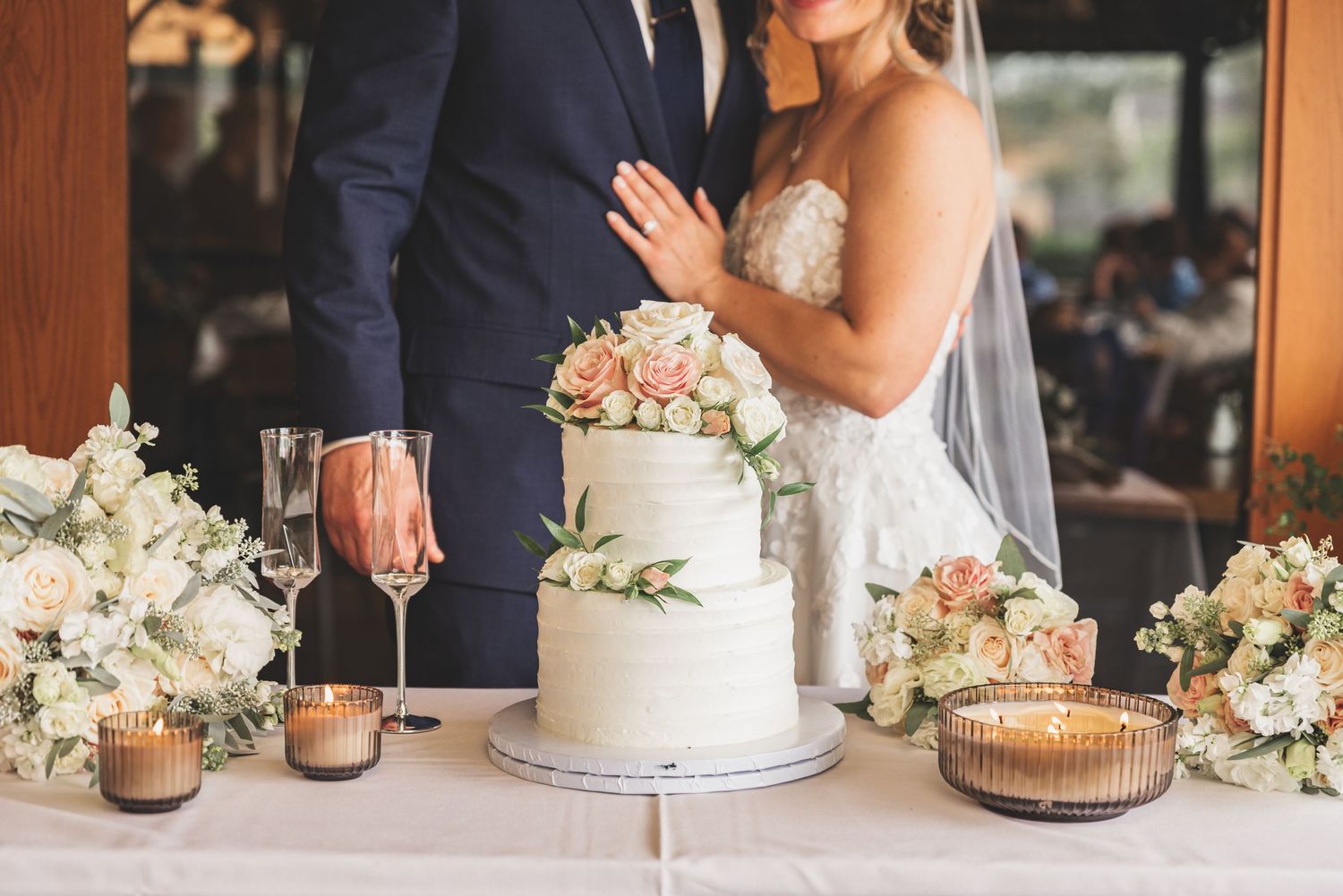Bride and groom cutting elegant two-tier white wedding cake adorned with blush roses and greenery at reception.