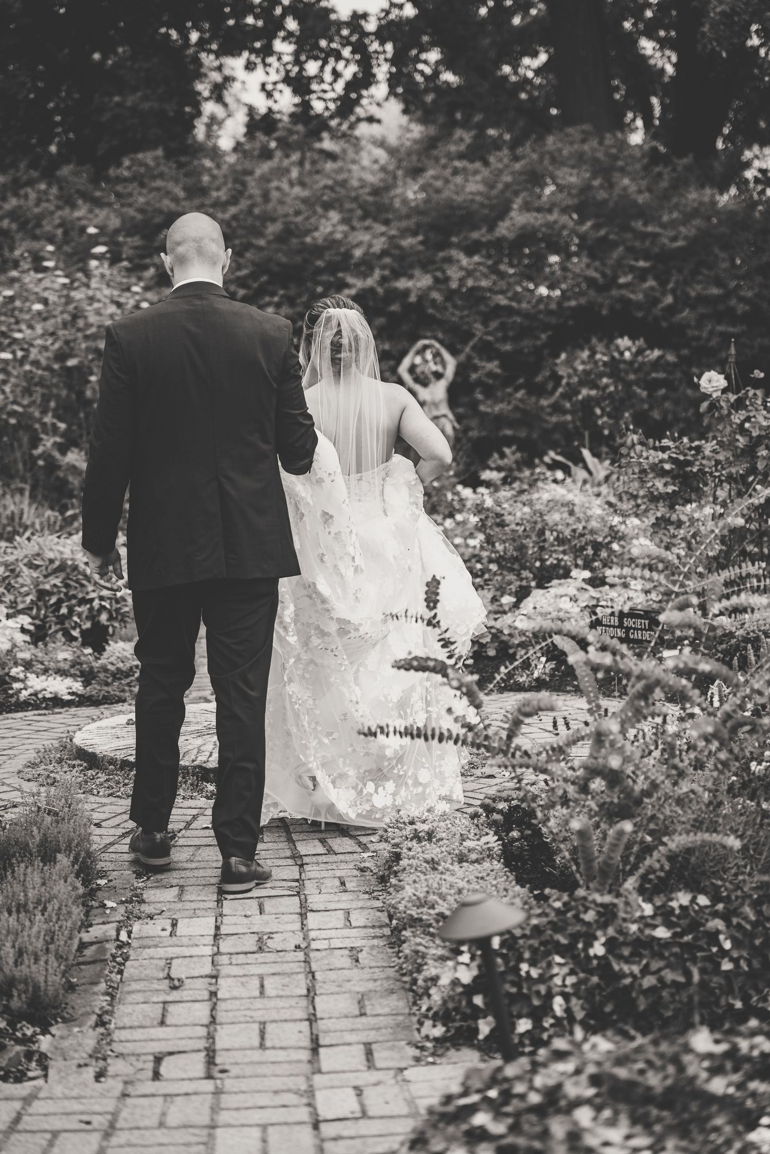 Black and white photo of bride and groom walking through lush garden pathway together.