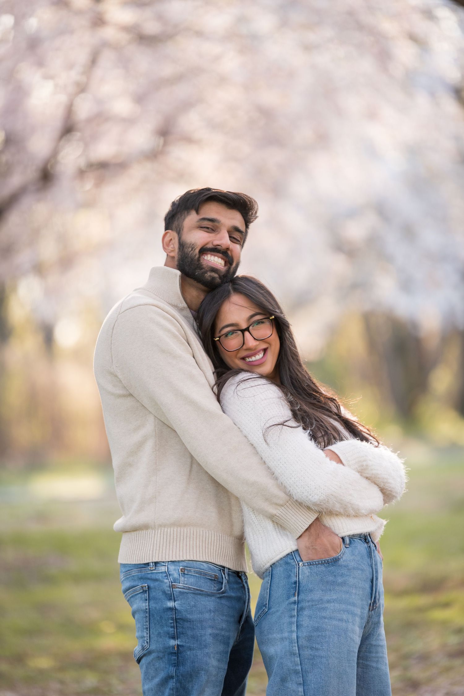 Happy couple embracing outdoors among spring cherry blossoms, wearing matching cream sweaters and blue jeans.