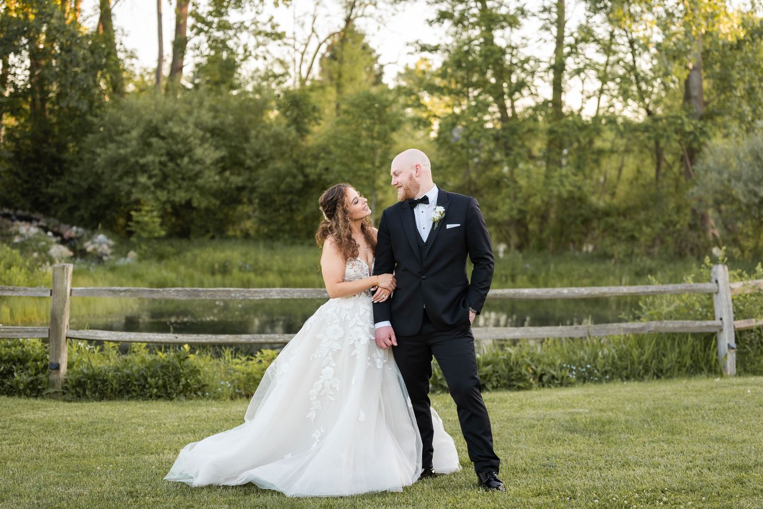 Bride and groom sharing a romantic moment near a wooden fence in a lush green outdoor wedding setting.