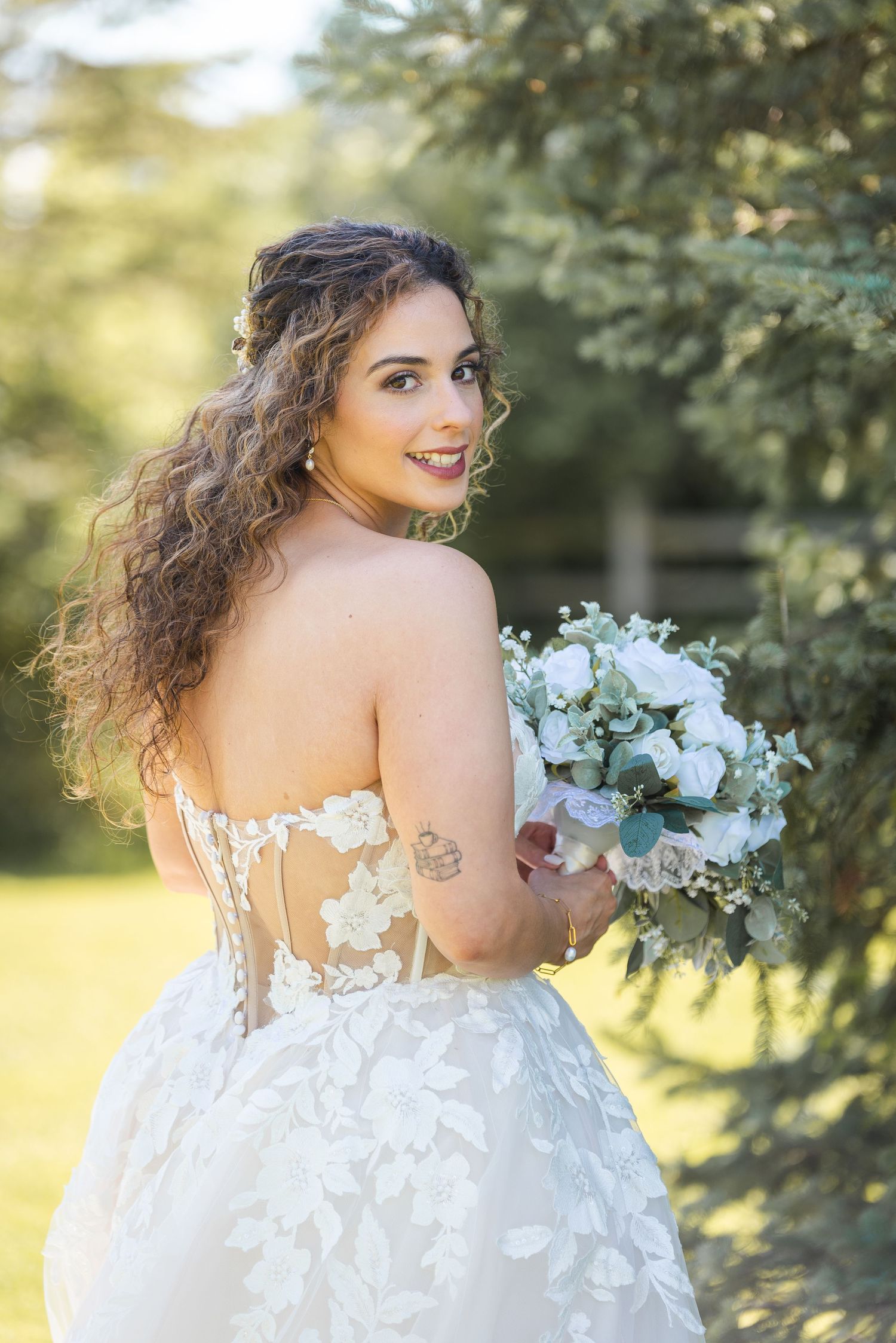 Bride in backless lace gown holding white bouquet smiling over shoulder.