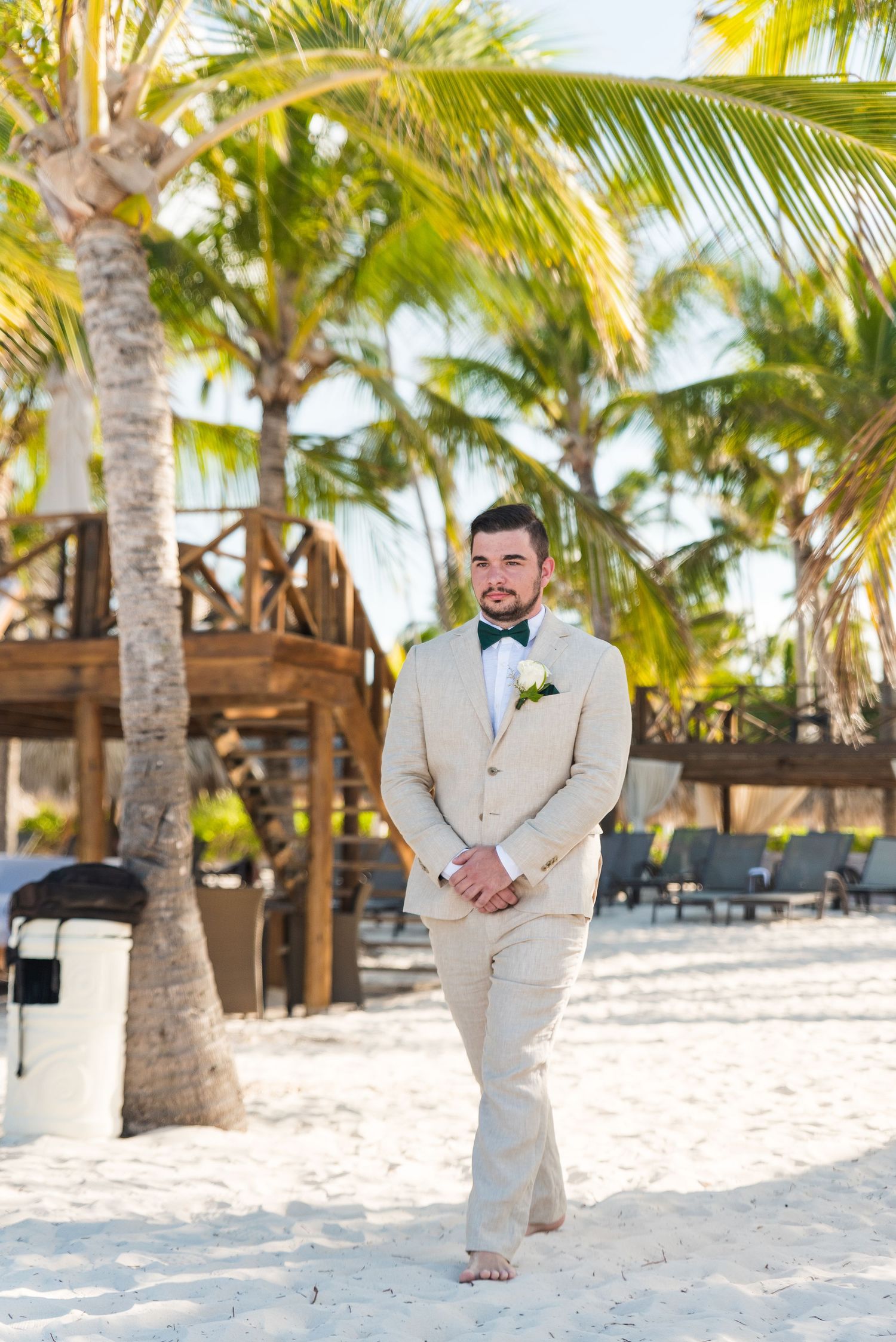 Groom in beige suit and green bow tie stands barefoot on white sand beach surrounded by palm trees.