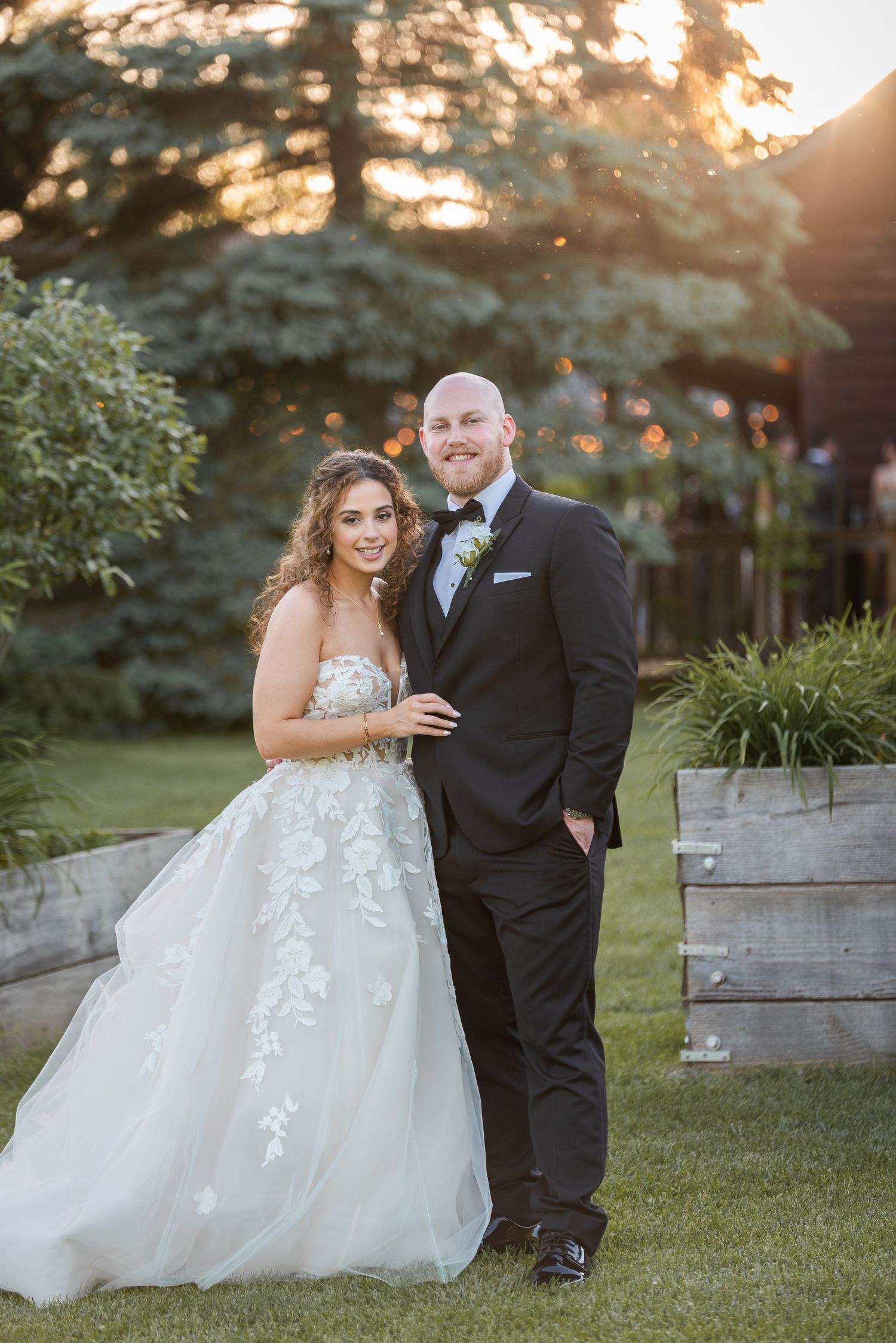 Bride in floral lace gown and groom in tuxedo posing together on lawn at golden hour sunset wedding.