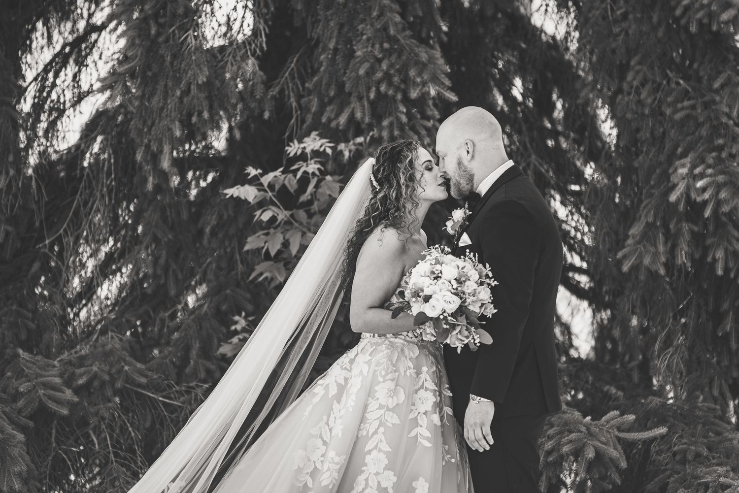 Black and white photo of bride and groom kissing under lush evergreen trees, bride holding bouquet.